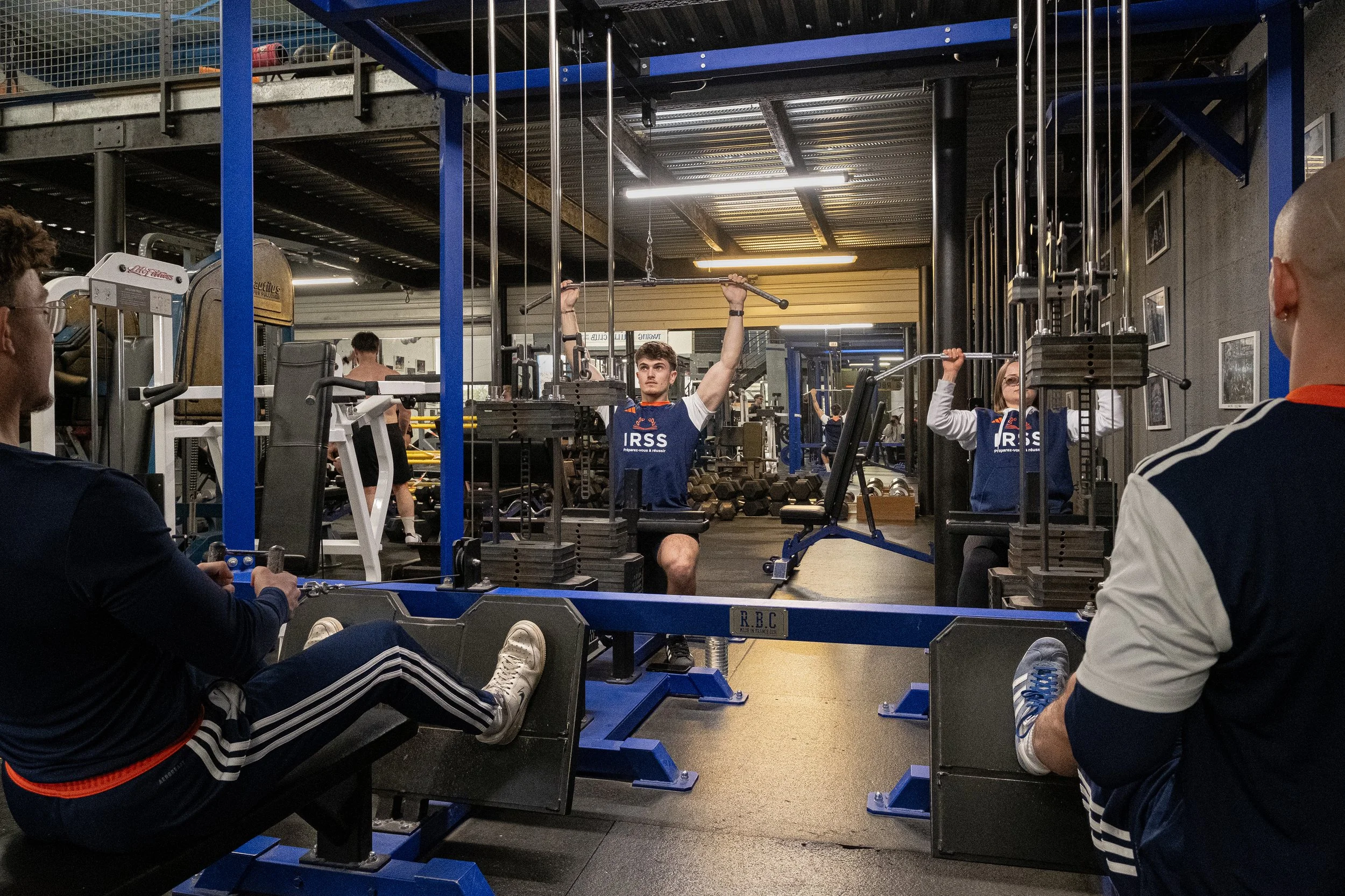 Homme de musculation faisant des exercices aux machines de musculation dans une salle de sport. Autres personnes s'entraînant en arrière-plan.