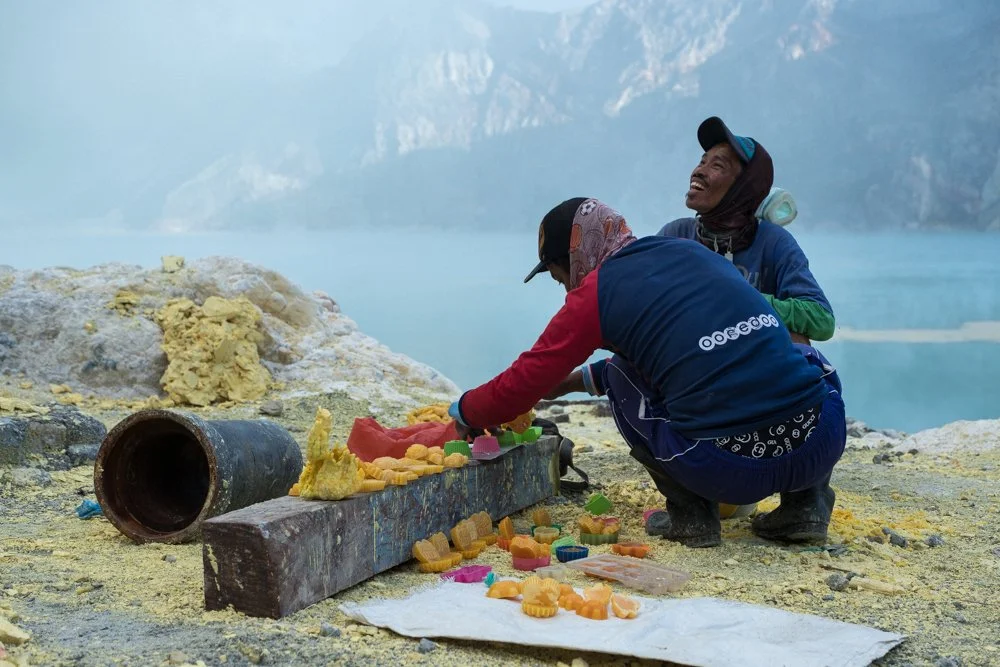 Deux personnes souriantes, habillées avec des vêtements de travail, manipulent des petits moulages de soufre coloré sur un terrain rocheux près d’un lac avec des falaises en arrière-plan.