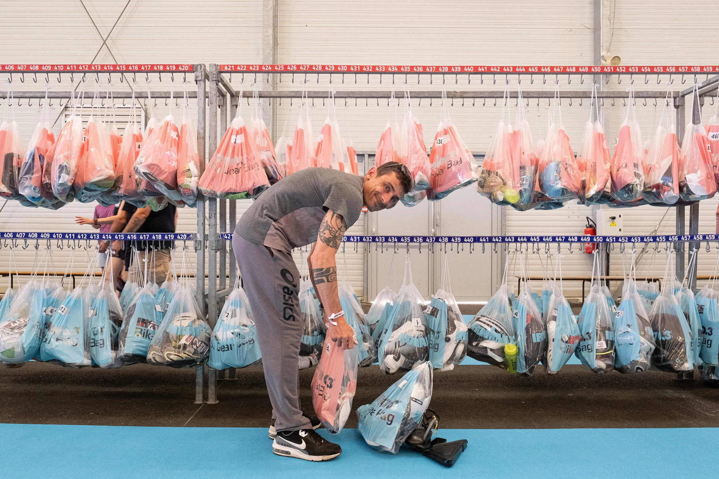 Un homme qui prépare ses affaires pour un triathlon, des rangées de sacs appartenant aux autres compétiteurs derrière lui.