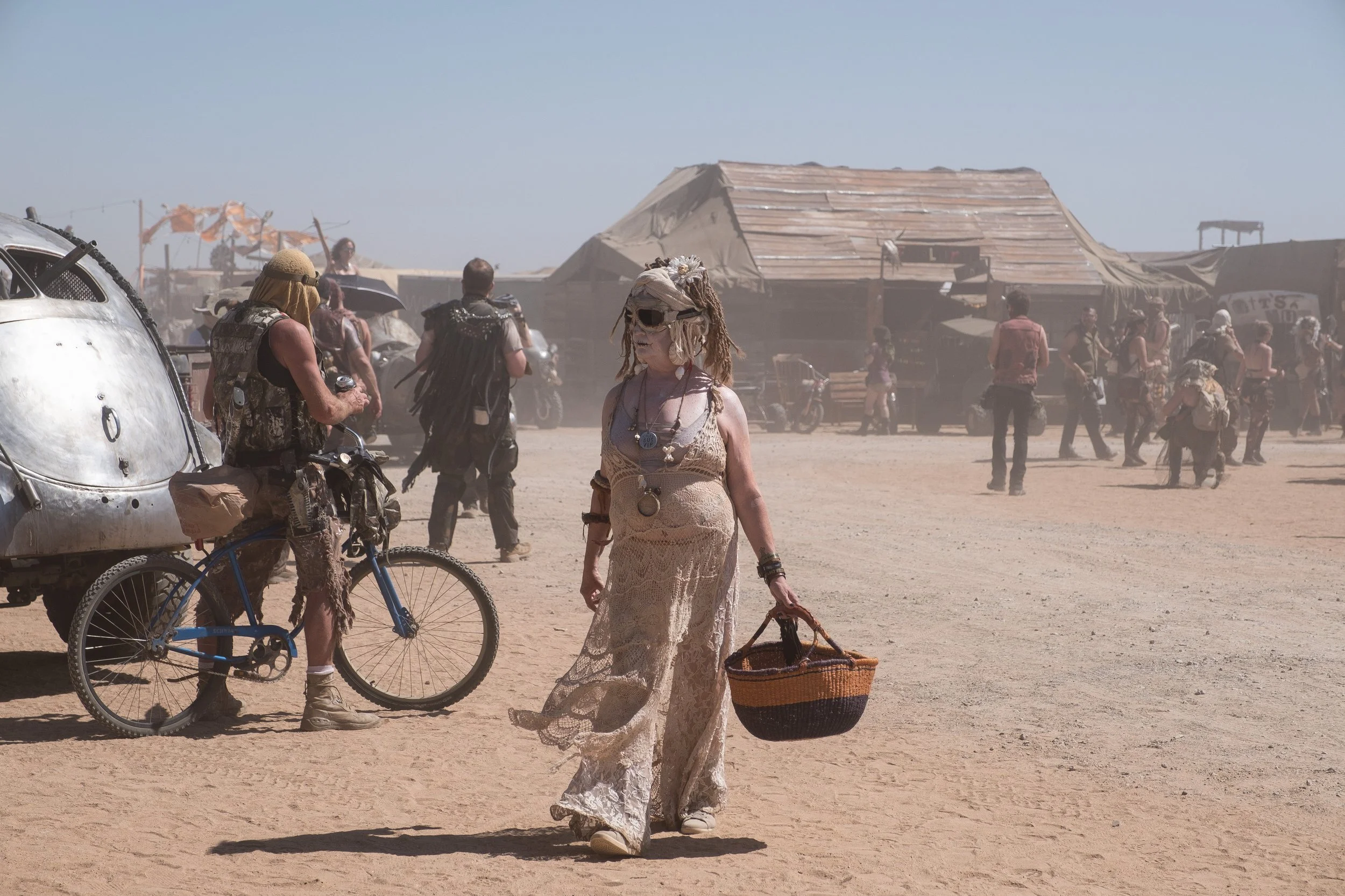 Une femme portant des vêtements en dentelle et des accessoires, portant un panier, marche dans un environnement poussiéreux avec des personnes et des structures en bois en arrière-plan, évoquant une scène de festival ou un rassemblement en plein air