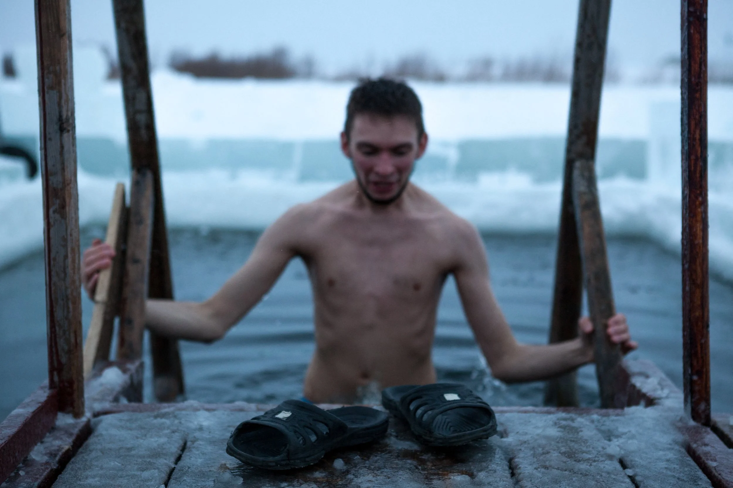 Jeune homme sortant d'une eau glacée dans un environnement enneigé, tenant une échelle en fer, avec une paire de sandales noires à l'avant.