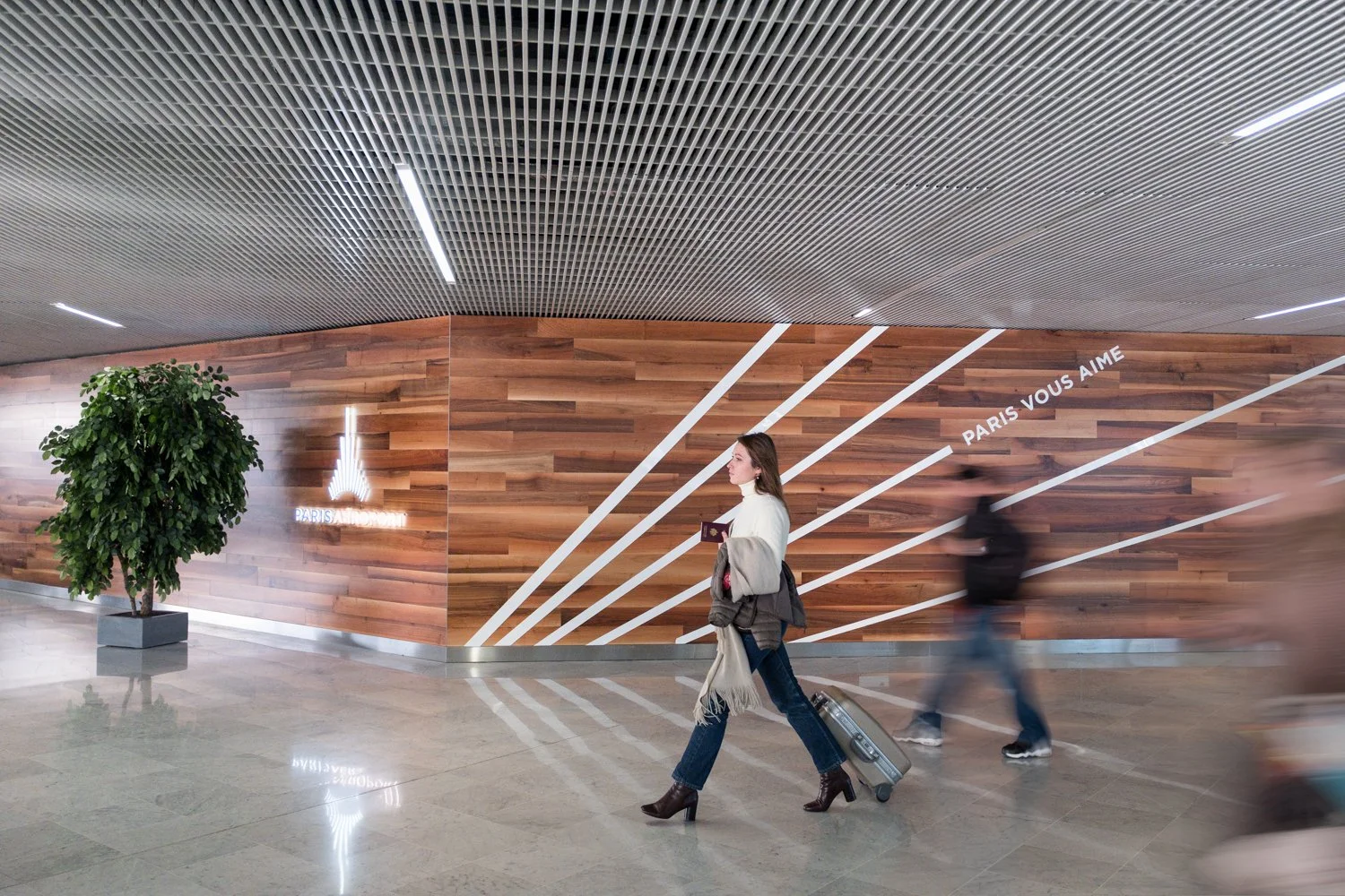 Une femme avec un bagage à roulettes marche devant un mur en bois avec des lignes blanches et la phrase 'PARIS VOUS AIME'. Il y a aussi un arbre en pot de chaque côté et un logo avec la Tour Eiffel sur le mur.
