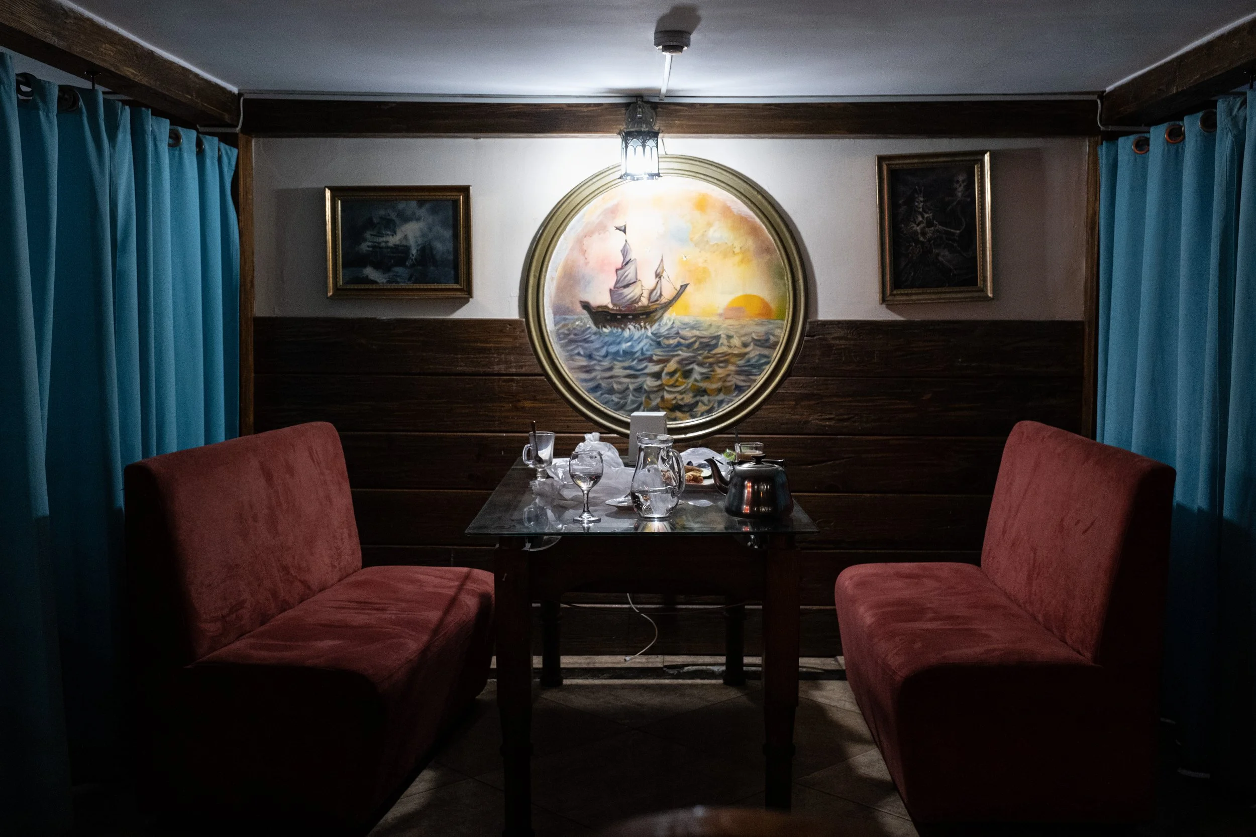 Une salle à manger intime avec deux banquettes rouges en velours de chaque côté d'une table en bois, décorée de verres et de carafes, un tableau rond représentant un bateau au coucher du soleil au centre et deux tableaux plus petits de chaque côté, r