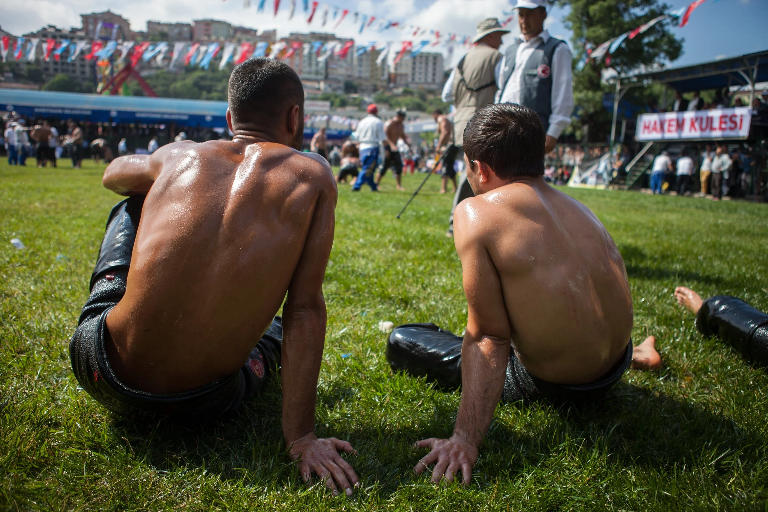 Deux jeunes lutteurs sont assis sur l'herbe, en culotte de cuir, lors d'une compétition de lutte en plein air, avec des fanions multicolores suspendus et beaucoup de personnes en arrière-plan.