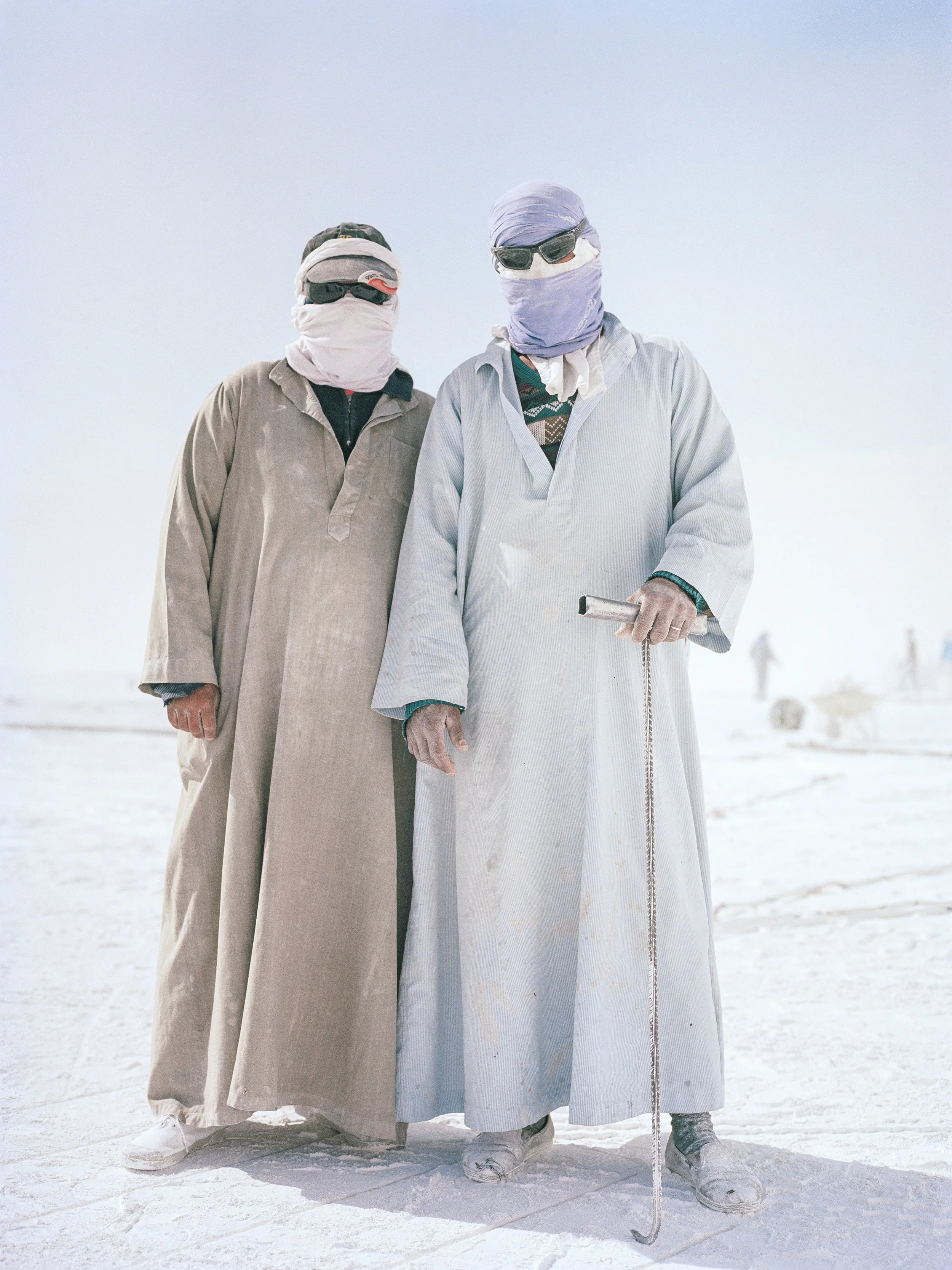 Deux hommes habillés en vêtements traditionnels et portant des lunettes de soleil, se tiennent dans un environnement blanc, l'un d'eux manipule un outil.