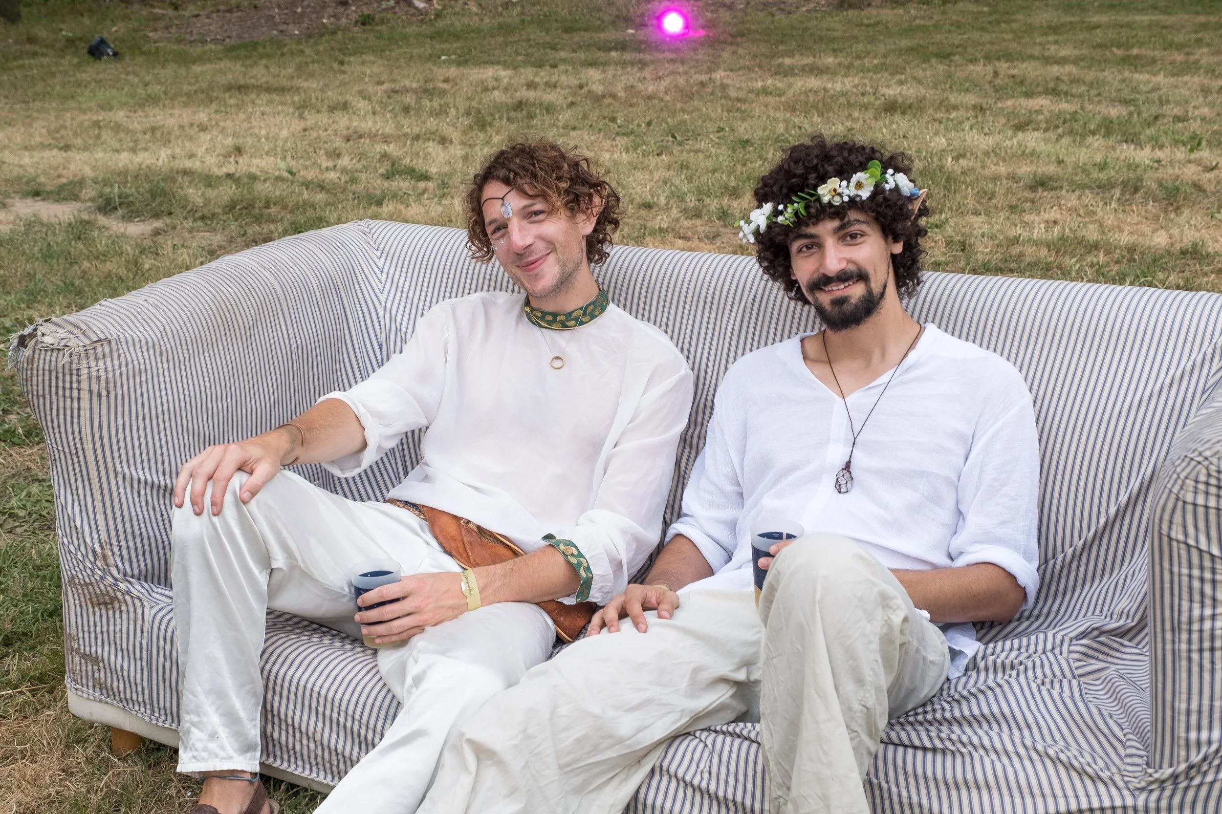 Deux hommes assis sur un canapé en extérieur, portant des vêtements blancs et décorés de couronnes de fleurs et d'accessoires de festival, souriant et détendus.