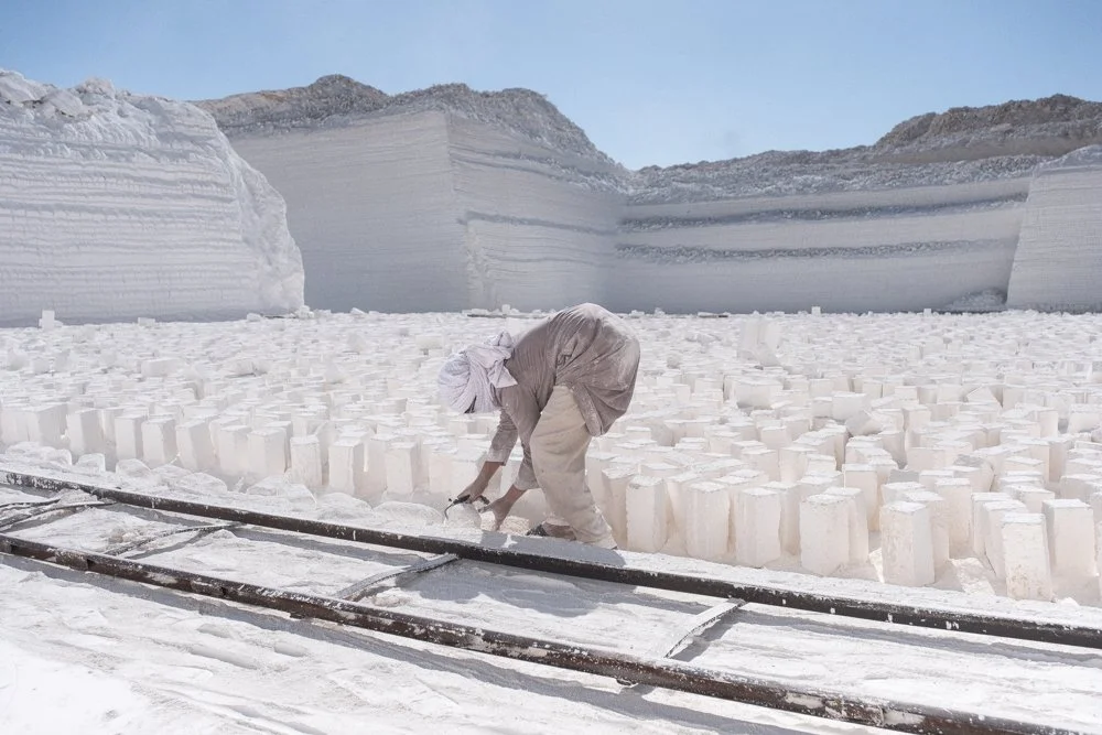 Un homme plié en deux met en position verticale des blocs de calcaire dans un paysage blanc avec des parois de calcaire en arrière-plan, sous un ciel clair.
