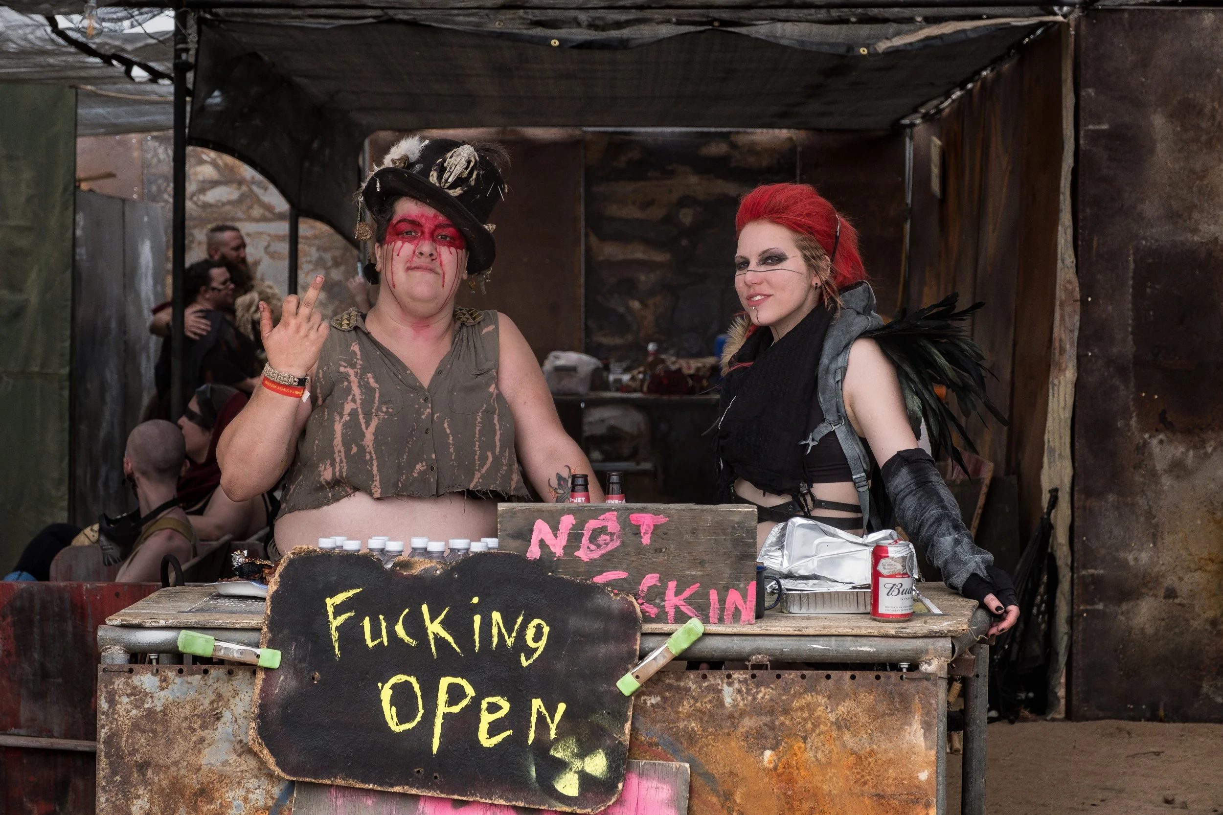 Deux femmes en costume post-apocalyptique dans un stand de fête, l'une fait un geste provocateur, l'autre sourit, avec des pancartes et des boissons sur le comptoir.