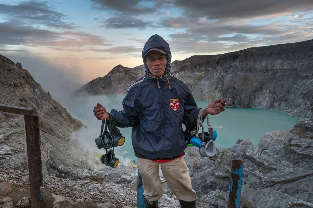 Homme portant une veste de pluie, tenant des masques à gaz ou des filtres à l'air, debout à côté d'un cratère volcanique avec un lac turquoise, dans un environnement brumeux et rocheux, au Kawah Ijen, Indonésie.