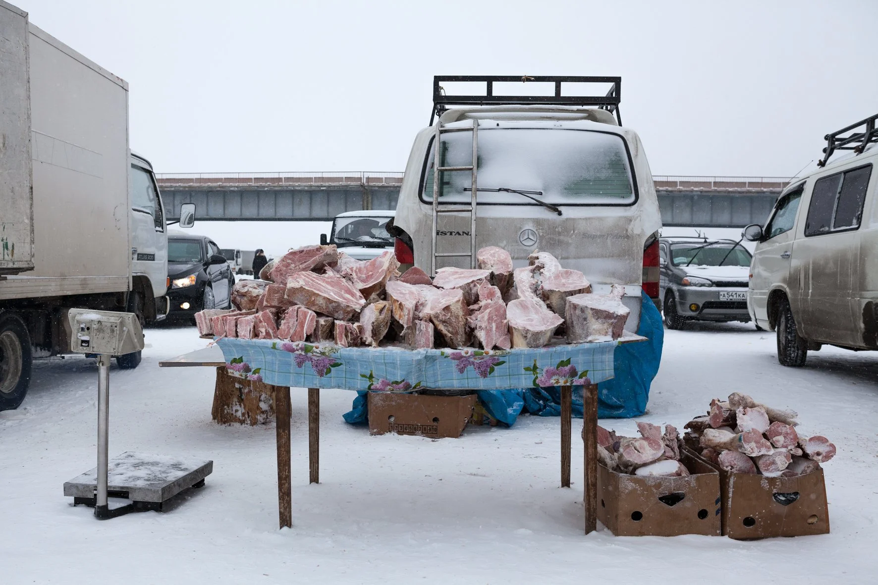 Vente de gros de morceaux de viande de porc, exposés sur une table en plein air, sous la neige, dans un parking avec des véhicules enneigés autour.