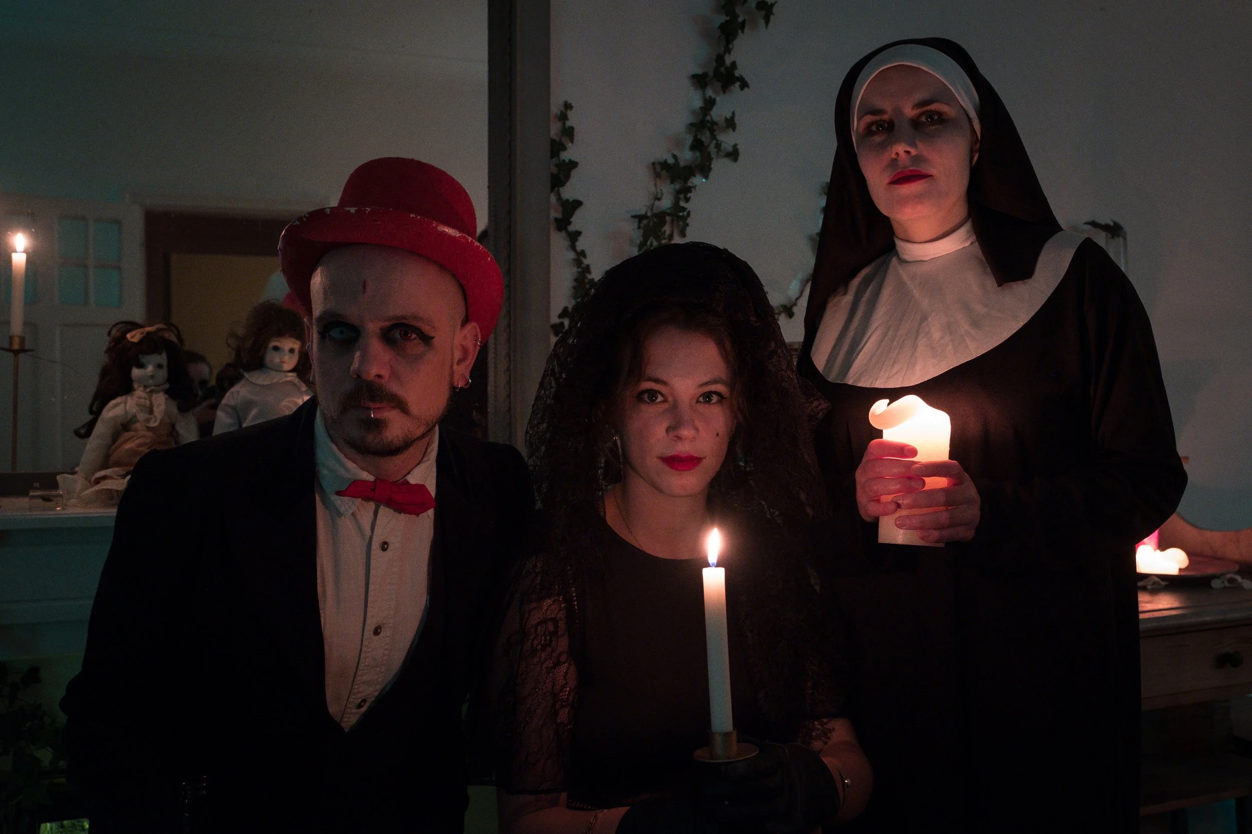 Trois personnes déguisées en personnages sinistres avec des bougies allumées dans une pièce sombre, décorée dans un style ancien et mystérieux.
