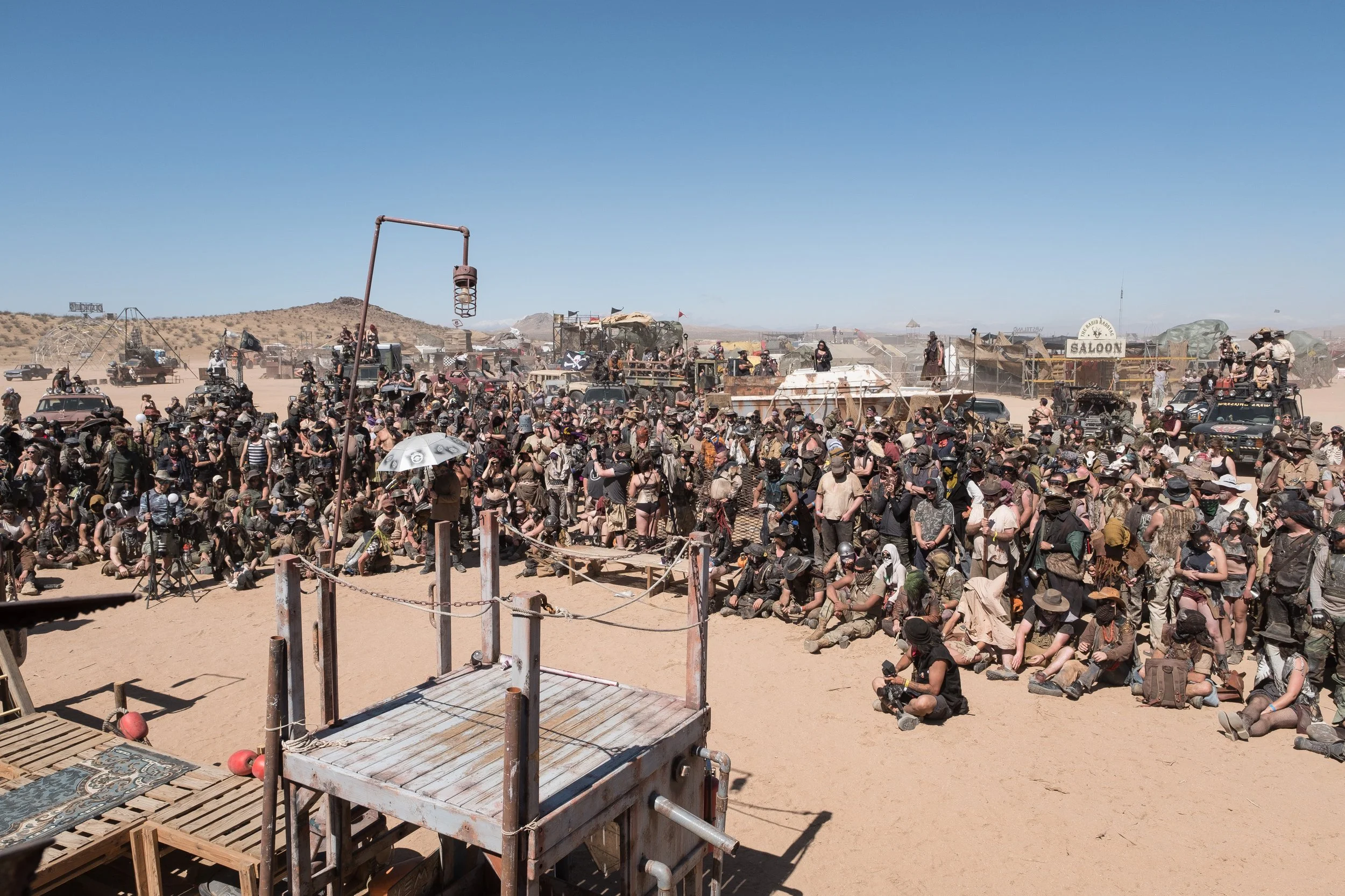 Rassemblement de personnes dans un désert, plusieurs portant des vêtements et accessoires de style post-apocalyptique, avec des véhicules et structures improvisées en arrière-plan, à Wasteland.