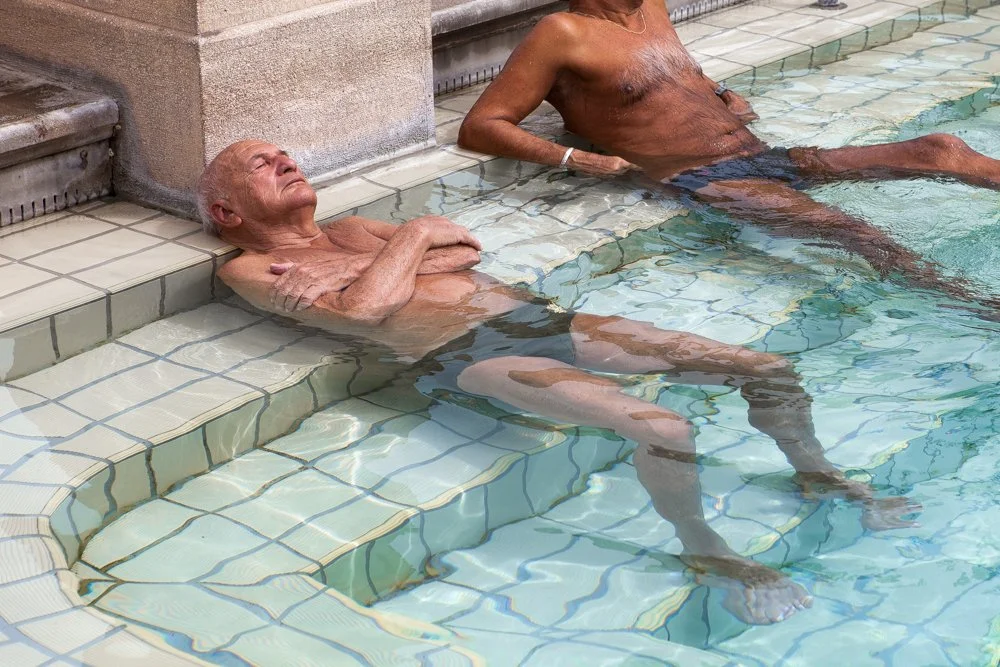 Deux hommes âgés se détendent dans une piscine, l'un assis sur le rebord avec les jambes dans l'eau, l'autre flottant sur le dos avec la tête contre le mur.