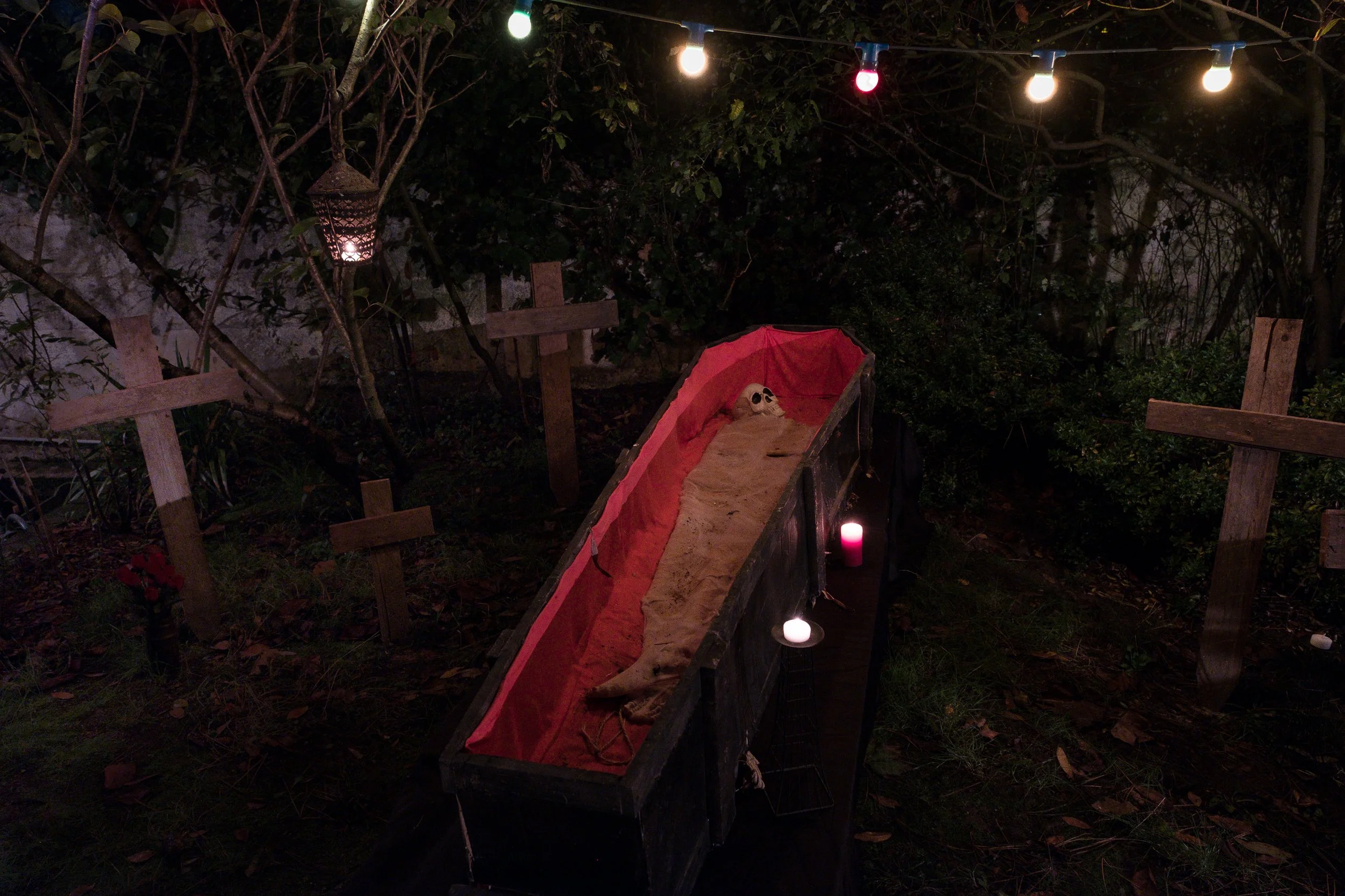 Un cercueil en bois noir avec une doublure rouge, contenant un squelette, dans un environnement nocturne décoré avec des lampes colorées et des bougies, entouré de crucifix en bois et de végétation pour fêter Halloween.