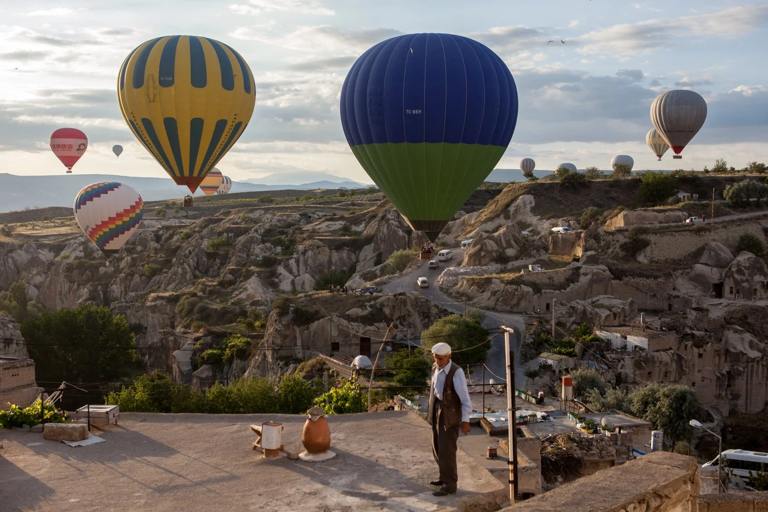 Plusieurs montgolfières colorées flottent dans le ciel au-dessus d'un paysage rocheux avec quelques habitations, un homme en tenue traditionnelle regarde depuis une terrasse en pierre au premier plan.