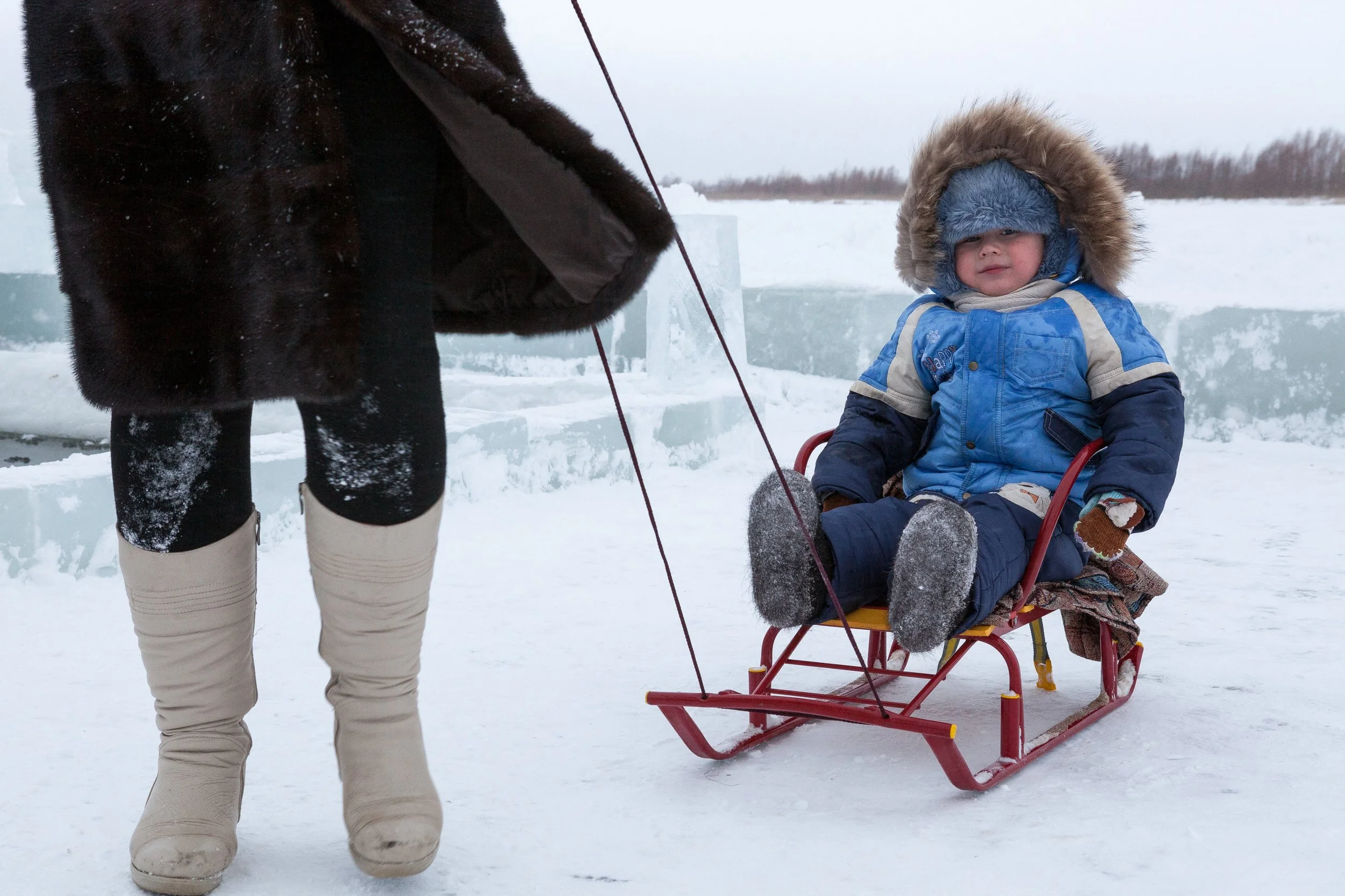 Enfant assis dans un traîneau dans un paysage enneigé, portant un manteau bleu avec une capuche en fourrure et un bonnet, en Russie.