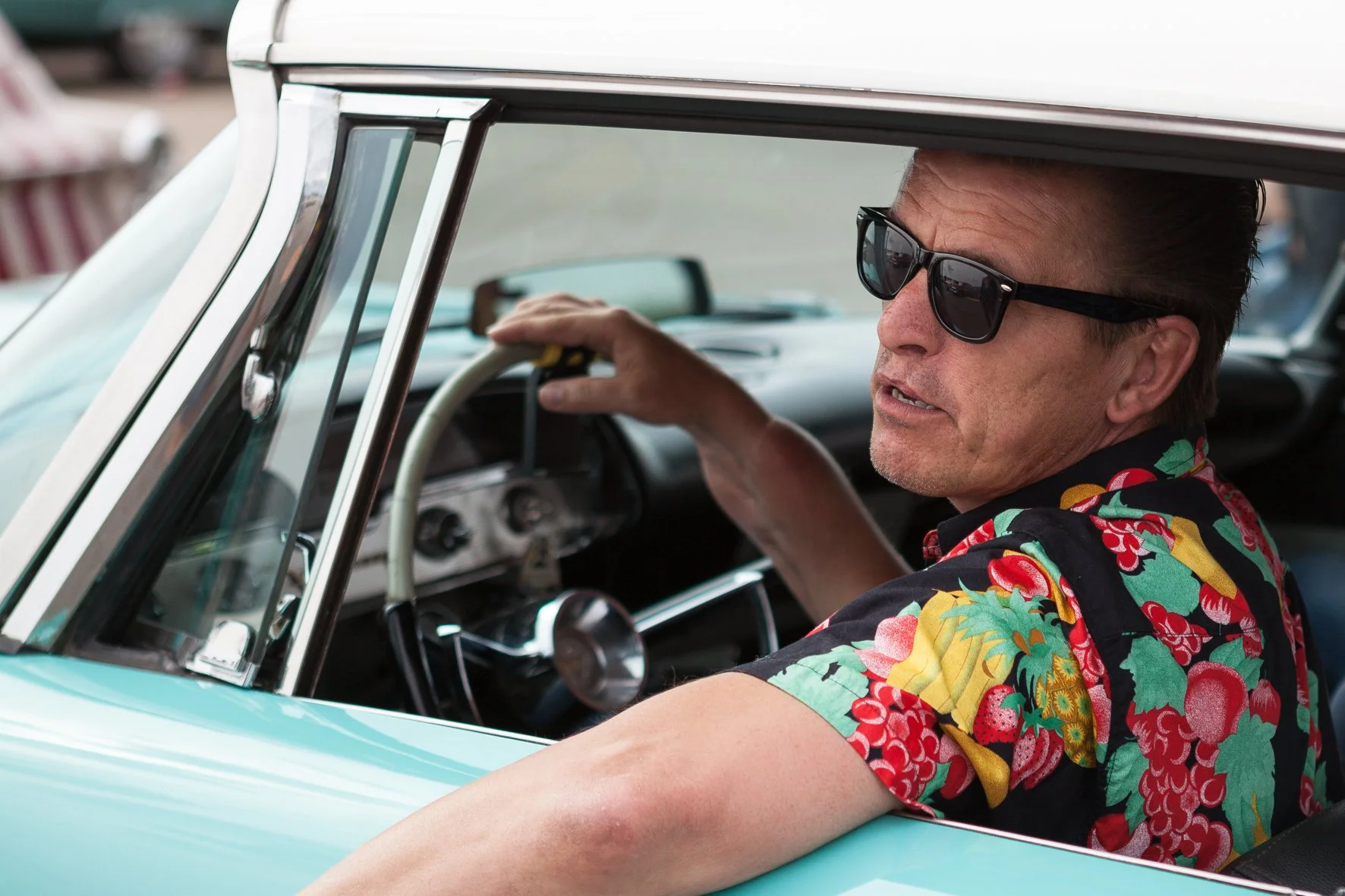 Un homme portant des lunettes de soleil et une chemise à motifs floraux colorés est assis dans une voiture vintage, tenant le volant.