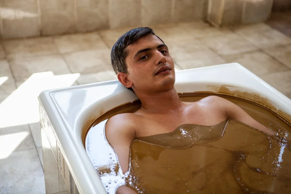 Homme allongé dans un bain thermal, entouré de pétrole, dans une pièce avec un sol en carrelage.