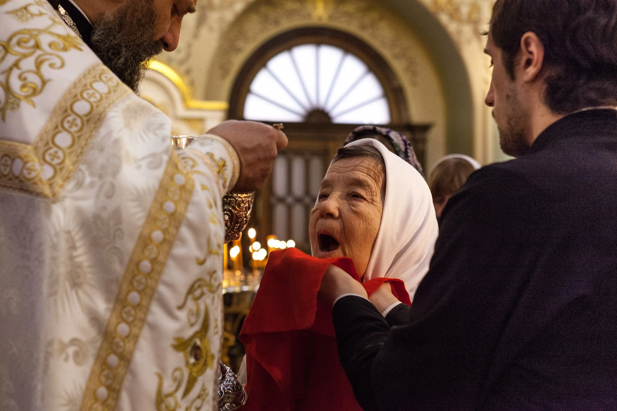 Une femme âgée, avec une tête couverte d'un foulard blanc, reçoit l'ostie lors d'une cérémonie religieuse dans une église, avec un prêtre en robe liturgique ornementée et un jeune homme en costume noir.