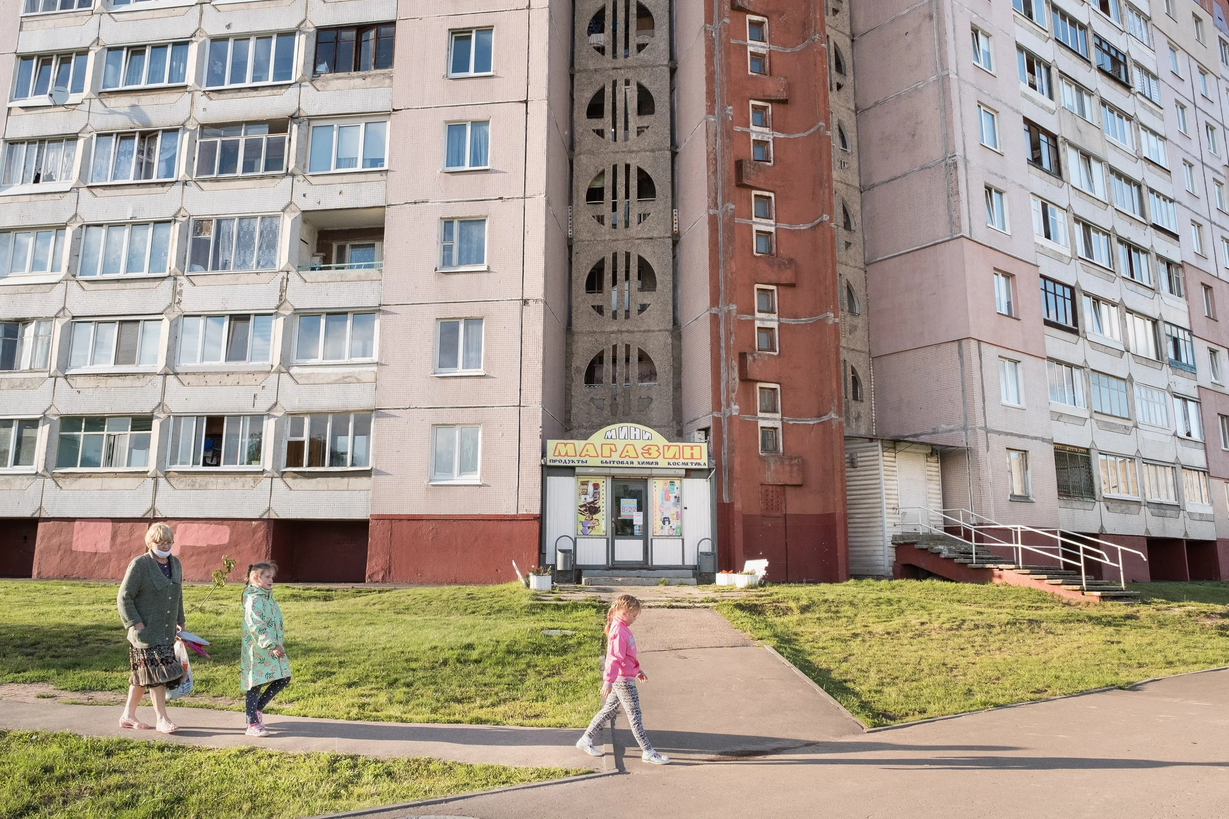 Une petite supérette au coin d'un immeuble résidentiel à plusieurs étages. Devant, trois personnes dont deux jeunes filles marchent sur un trottoir en herbe.