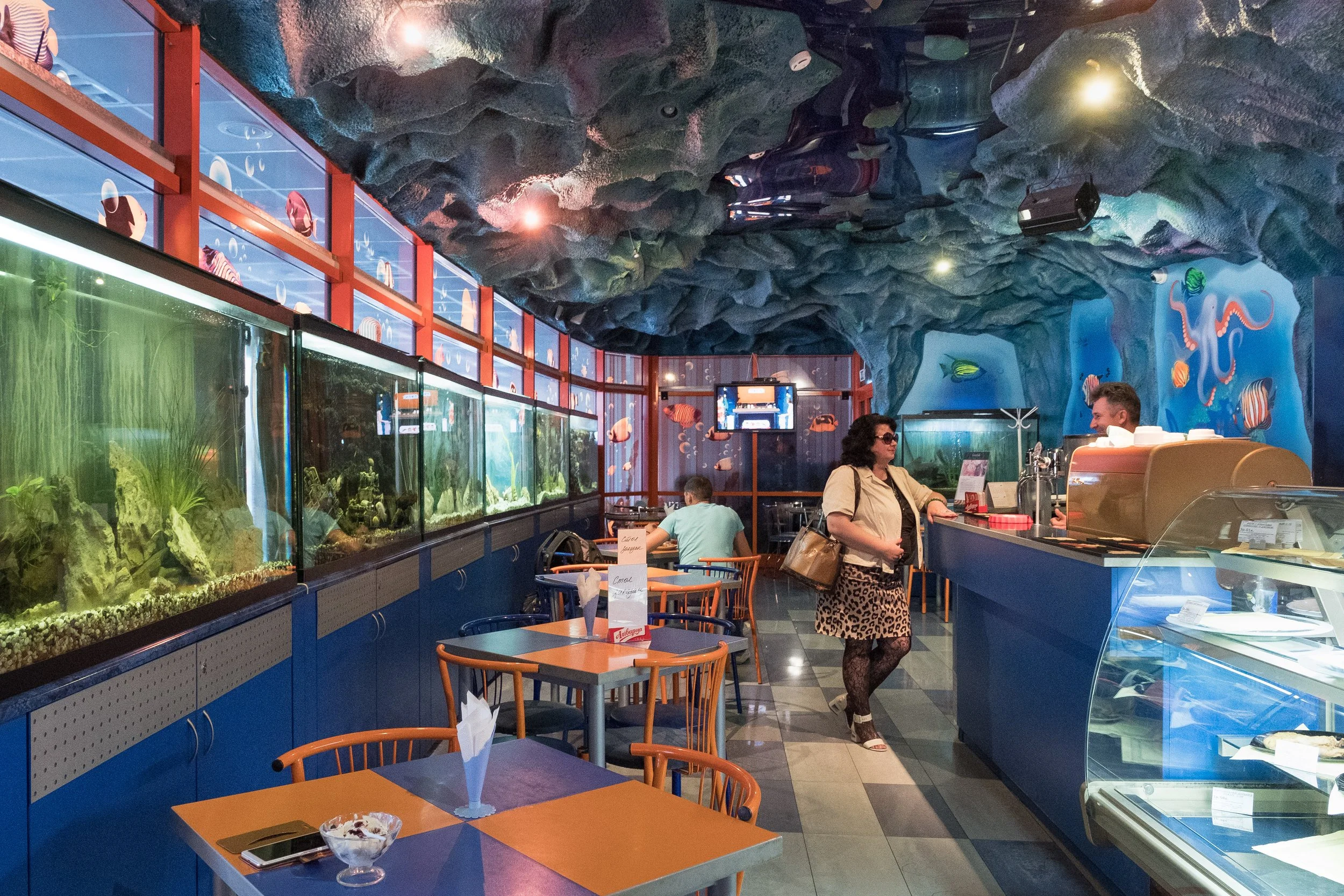 Intérieur d'un restaurant avec mur et plafond aménagés en thème marin, avec aquariums et décor sous-marin, tables et chaises en bois, un comptoir de service, une femme avec un sac et un homme derrière le comptoir dans la gare de Minsk.