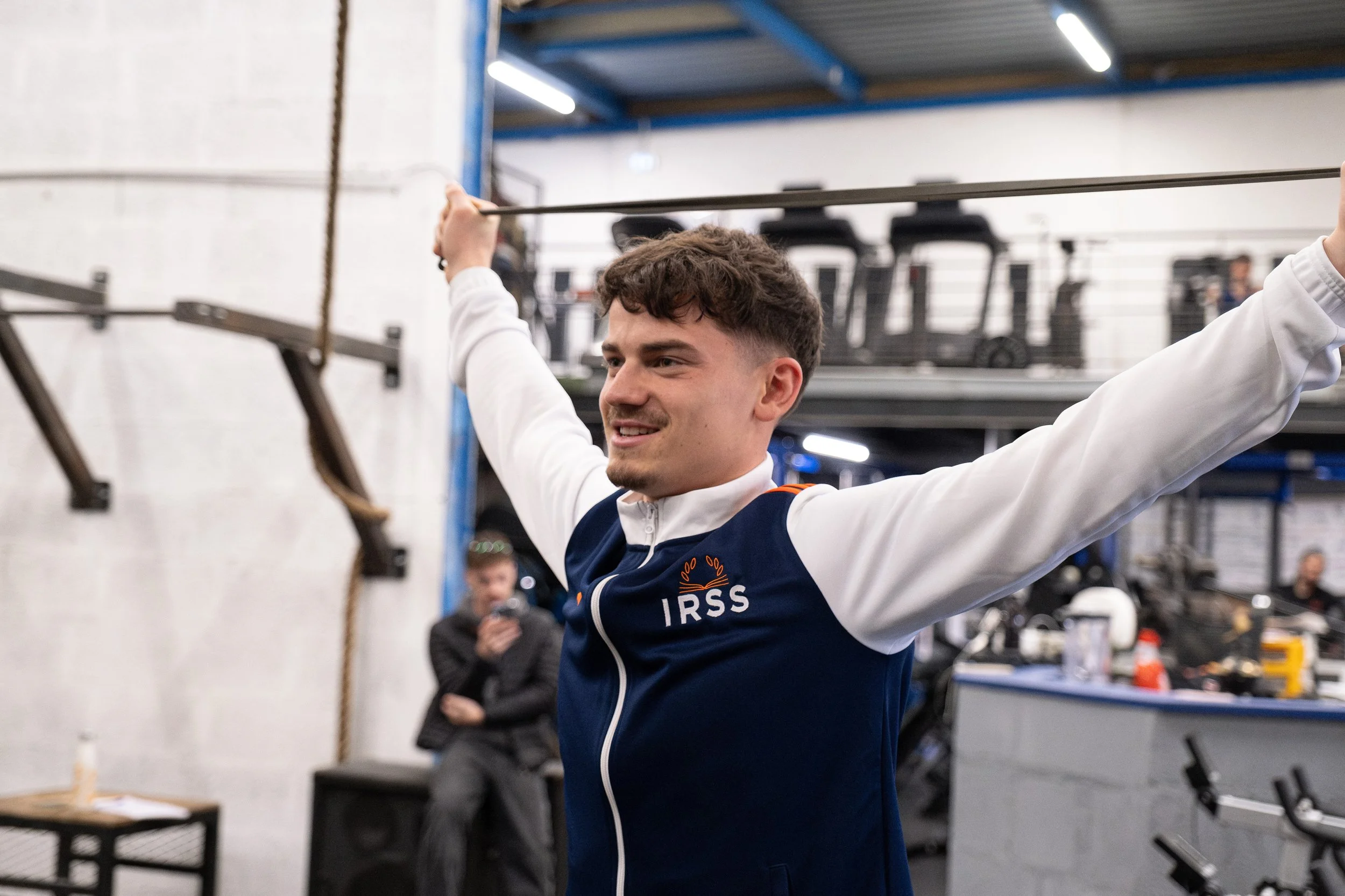 Un jeune homme dans une salle de sport, portant un sweat à capuche avec la mention IRSS, étire ses bras avec une barre. En arrière-plan, une personne est assise et regarde son téléphone.