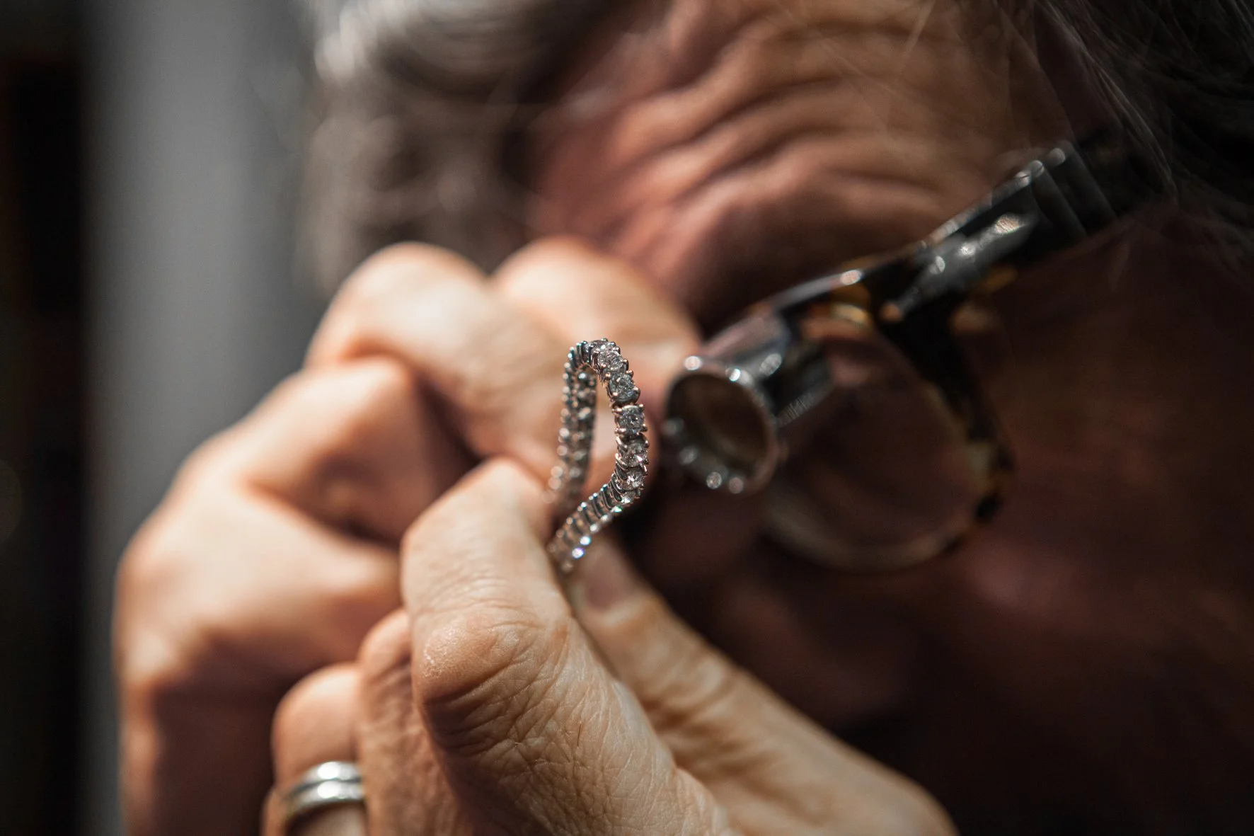 Une personne portant des lunettes, regarde avec une loupe un bracelet orné de diamants dans sa main. La personne porte également plusieurs anneaux, une montre et a les cheveux gris.