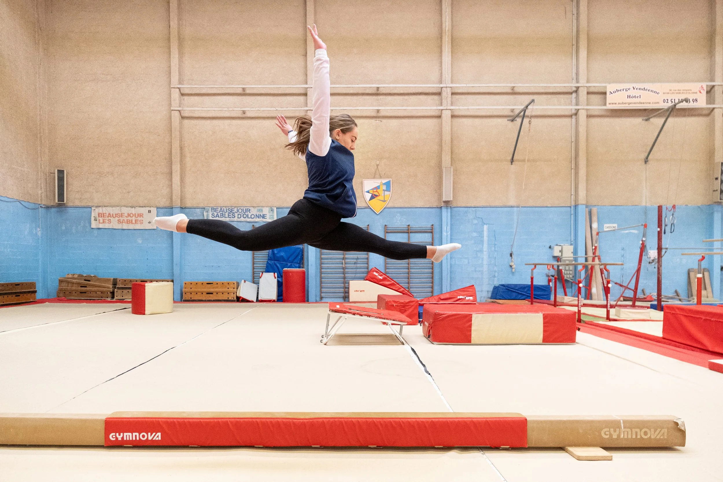 Une gymnaste effectuant une figure en saut dans un gymnase, avec un saut en étoiles, dans un environnement d'entraînement avec des équipements de gymnastique.