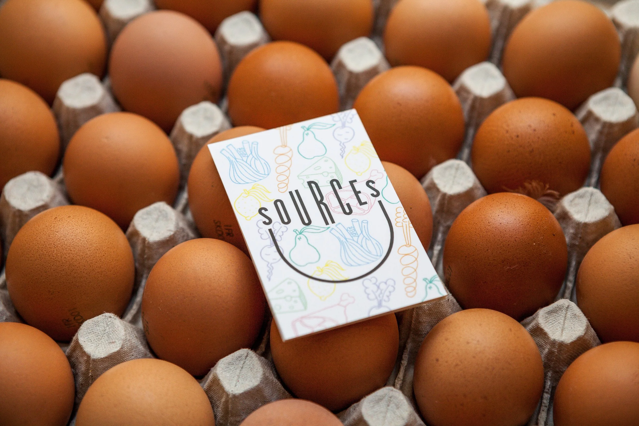 Une boîte contenant des œufs bruns, avec une carte de visite portant le mot 'SOURCES' et des dessins de légumes colorés.