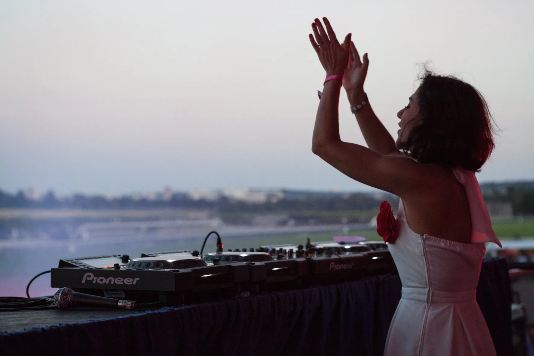 Une DJ femme en robe blanche avec les bras levés, près de sa table de DJ avec des équipements Pioneer, avec l'hippodrome de Longchamp en arrière-plan au crépuscule.