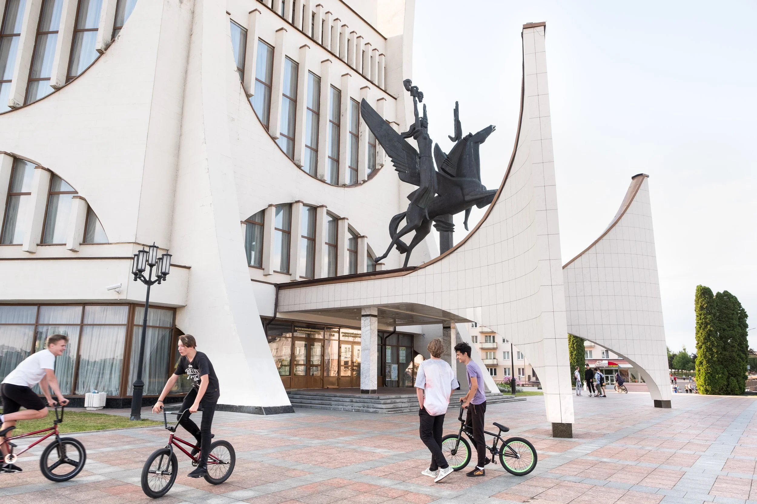 Un théatre moderne blanc en Biélorussie avec une sculpture en haut représentant un cheval ailé avec un personnage tenant une torche, et plusieurs jeunes adultes avec des BMX devant.