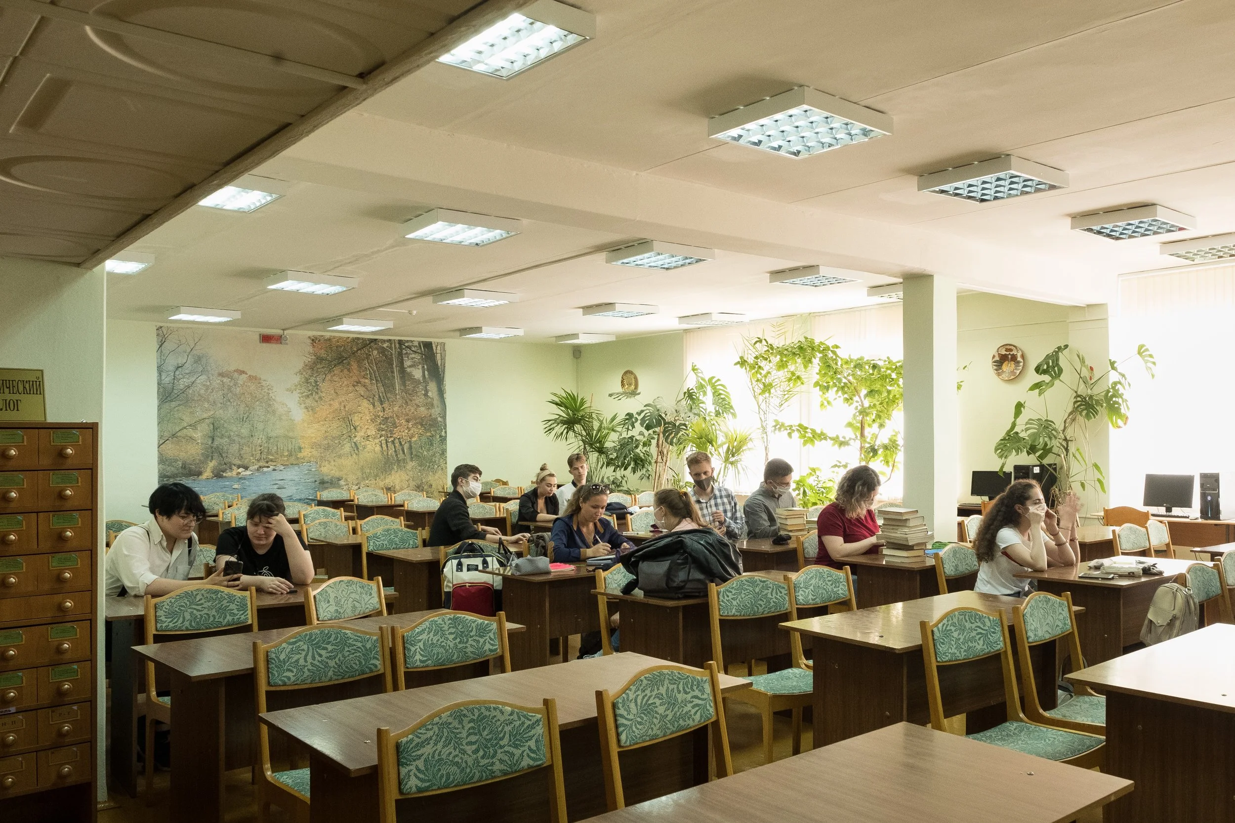 Salle de cours avec plusieurs étudiants assis à des tables, certains utilisant des ordinateurs ou lisant des livres, dans un environnement lumineux avec des plantes vertes et une grande peinture murale de nature à Gomel, en Biélorussie.