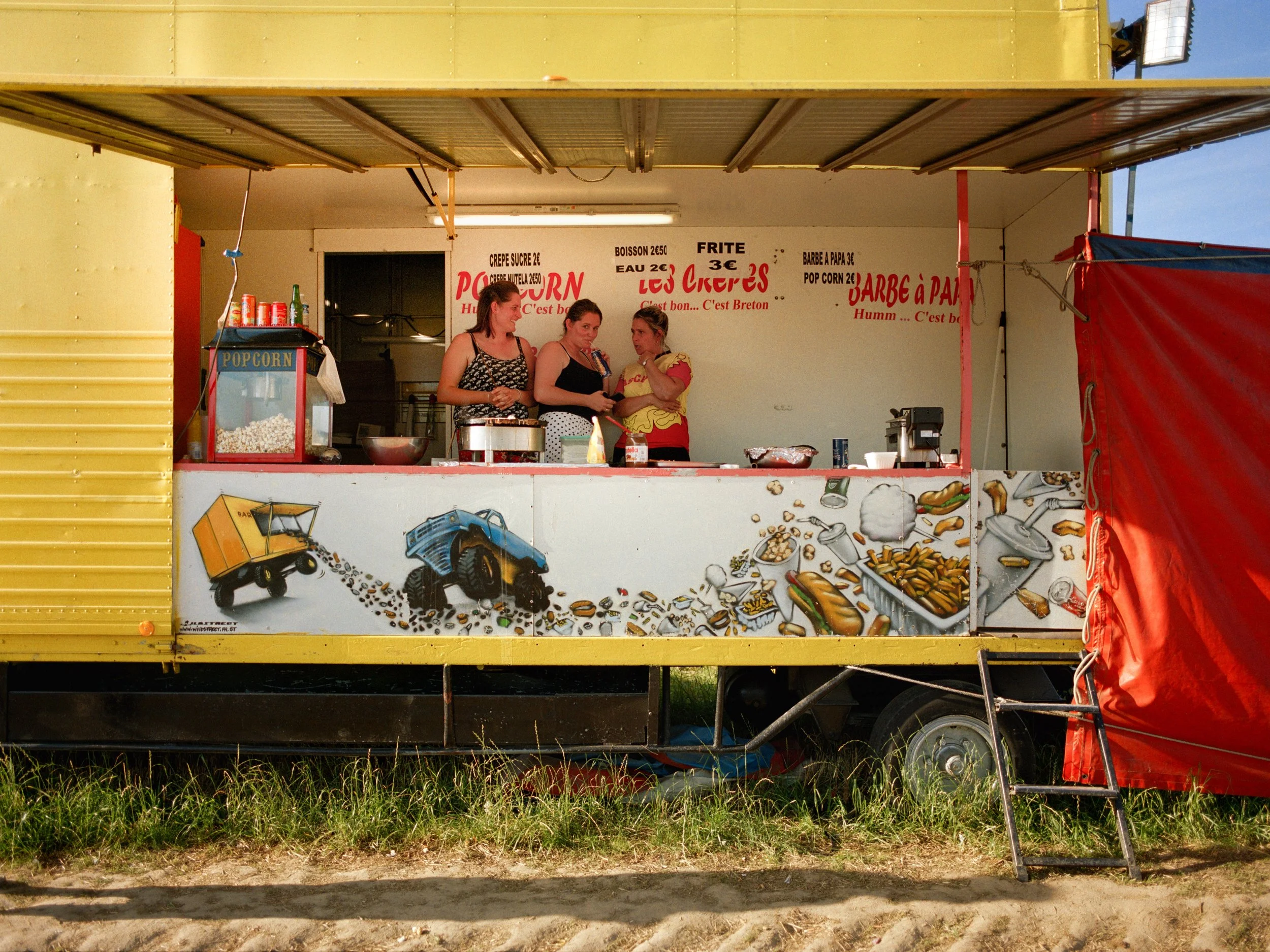 Stand de nourriture avec trois femmes derrière le comptoir, décor de dessins de camions et de nourriture en bas, menu écrit à l'arrière. Lignes de prix pour popcorn, crêpes, boissons, frites, barbes à papa, et autres spécialités bretonnes.