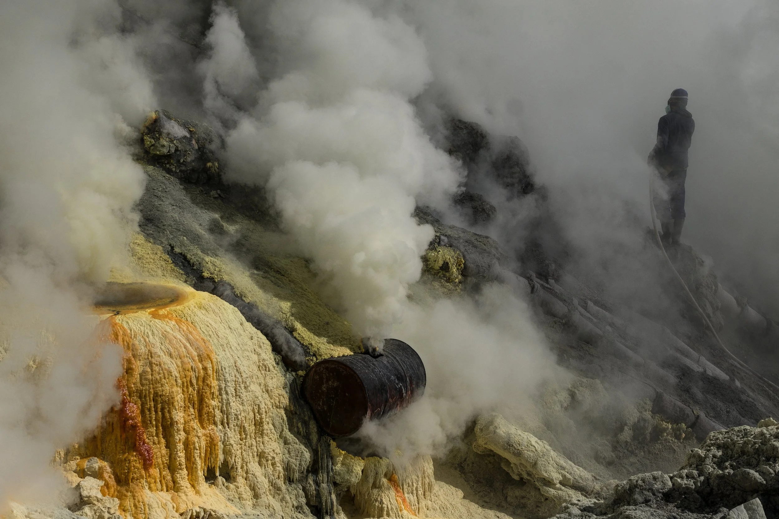 Une scène géothermique avec de la vapeur s'échappant de fumerolles, des dépôts minéraux jaunes et oranges, un tuyau rouillé, et une personne debout portant un casque et se tenant sur des rochers dans un environnement volcanique.