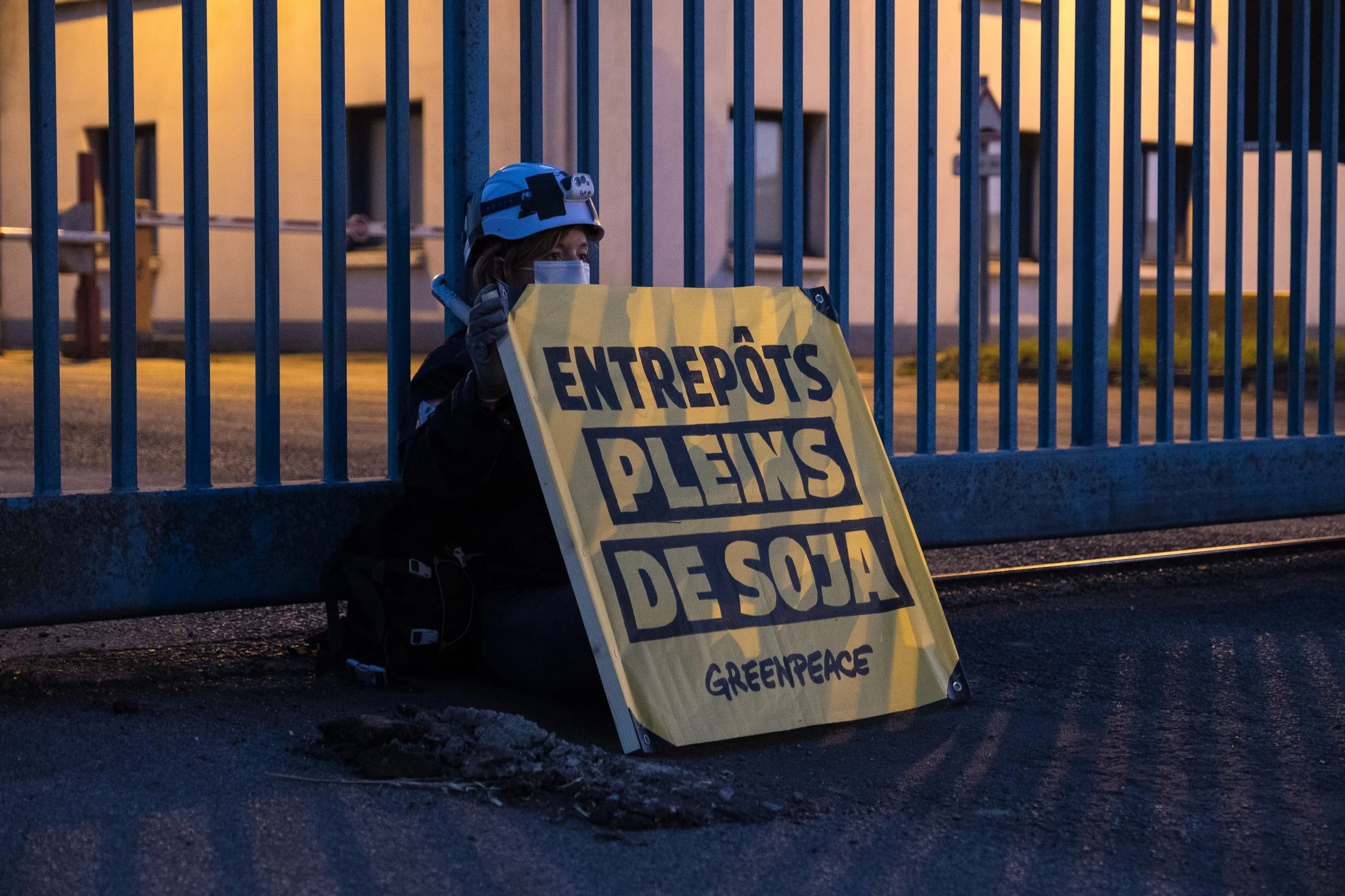 Une activiste de Greenpeace assise au sol tient une pancarte jaune indiquant 'ENTREPÔTS PLEINS DE SOJA'.