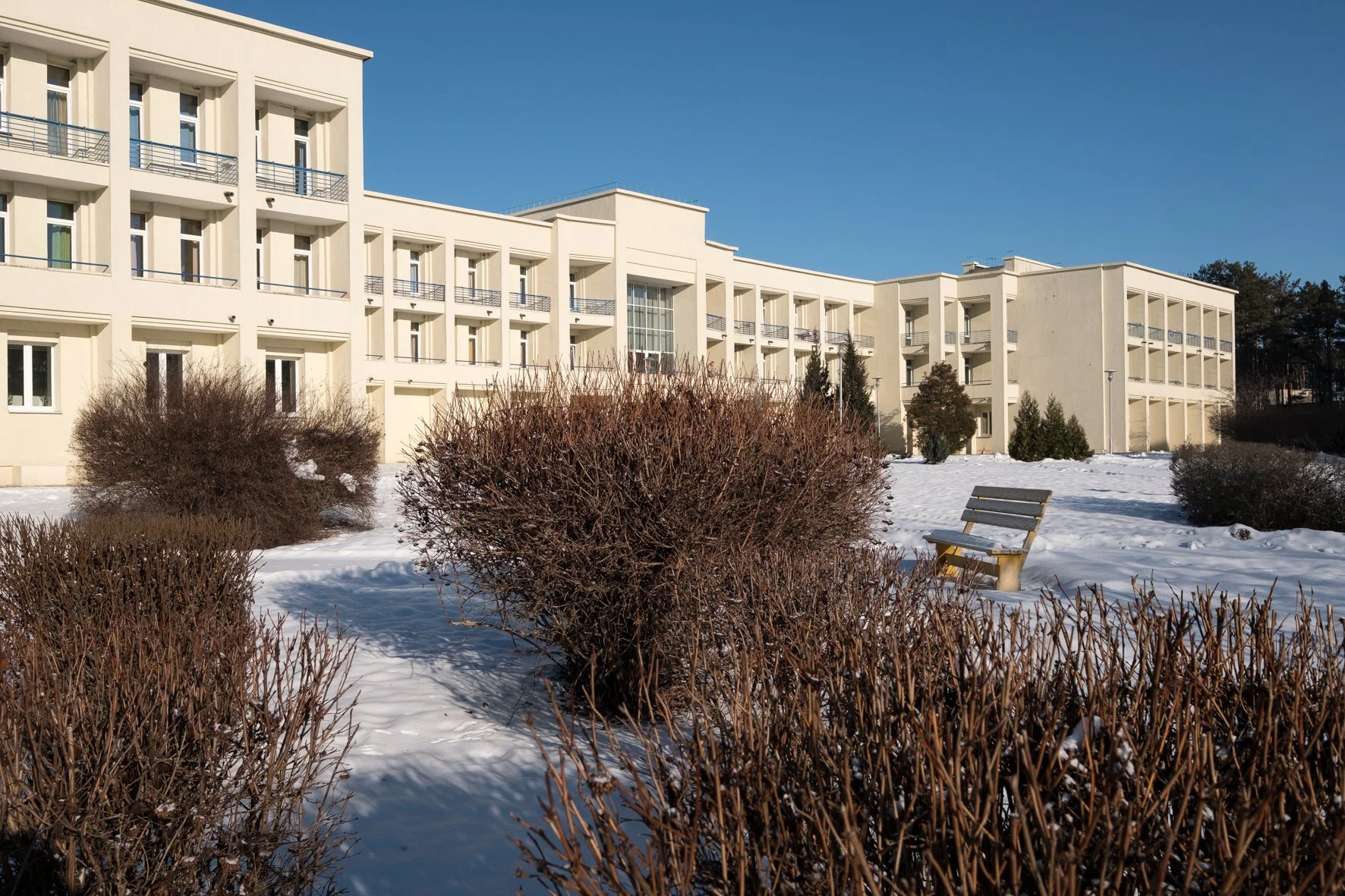 Un immeuble moderne blanc avec plusieurs étages et balcons, sous un ciel bleu clair, avec des buissons et de la neige au sol, ainsi qu’un banc en bois dans un espace extérieur.