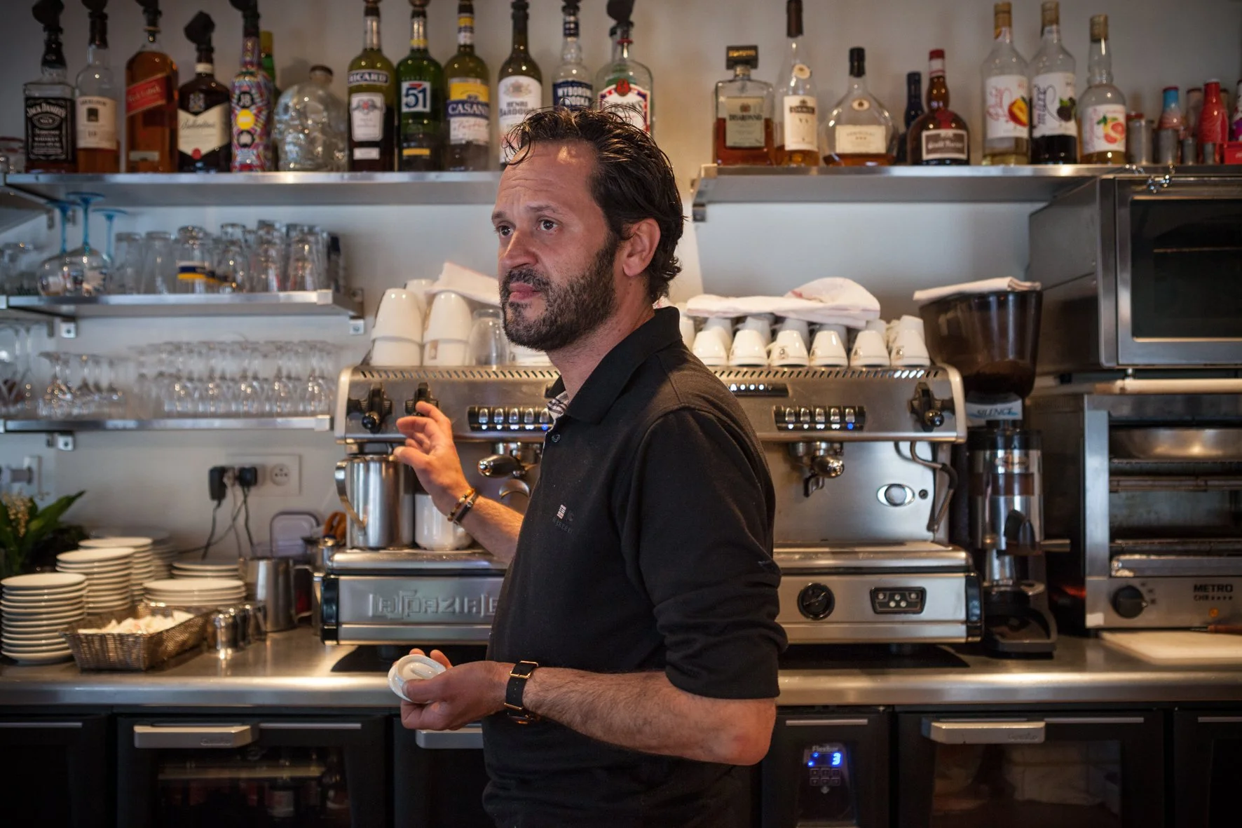 Un homme dans un café, en train de préparer un café avec une machine à café professionnelle. Le bar est en arrière-plan, avec des bouteilles d'alcool, des tasses, et du matériel de cuisine visible.
