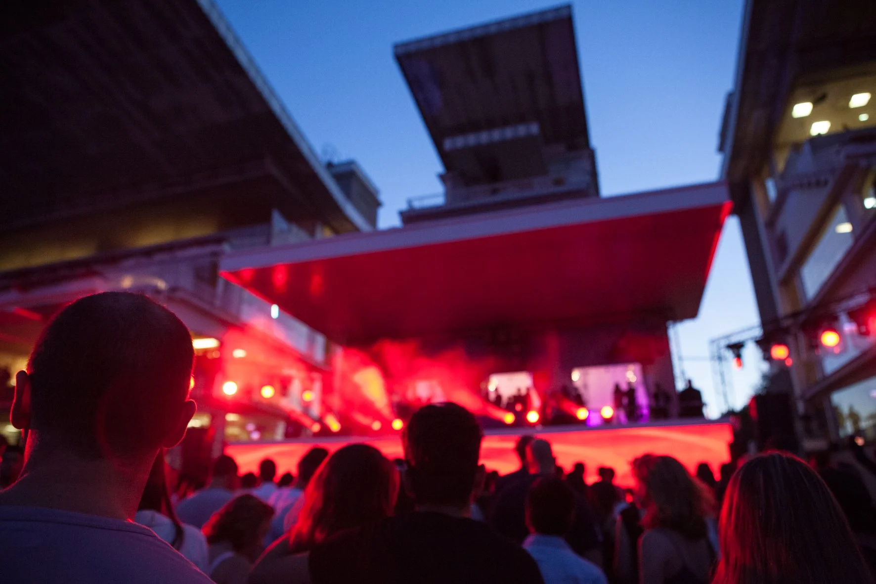 Un DJ set en plein air à l'hippodrome de Longchamp avec une scène éclairée en rouge, une foule de spectateurs regardant le spectacle, à la tombé du jour.