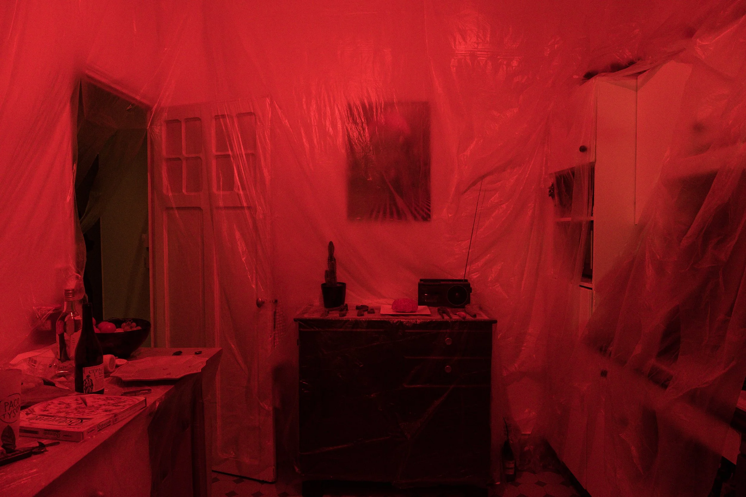 Une pièce d'intérieur recouverte de plastique rouge, avec une étagère à gauche, un meuble central avec une plante et un radio, une porte à gauche, et une affiche ou tableau au mur.