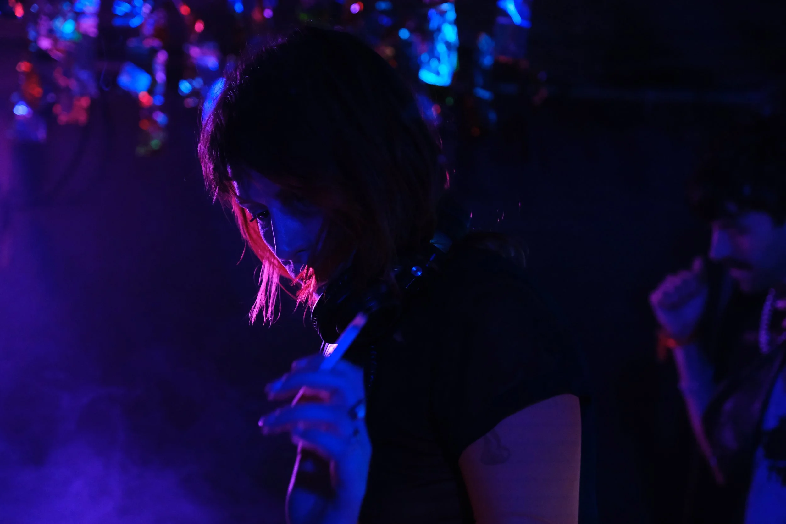 Une femme avec des cheveux bruns qui tient une cigarette dans un environnement sombre éclairé par des lumières colorées, avec une autre personne en arrière-plan.