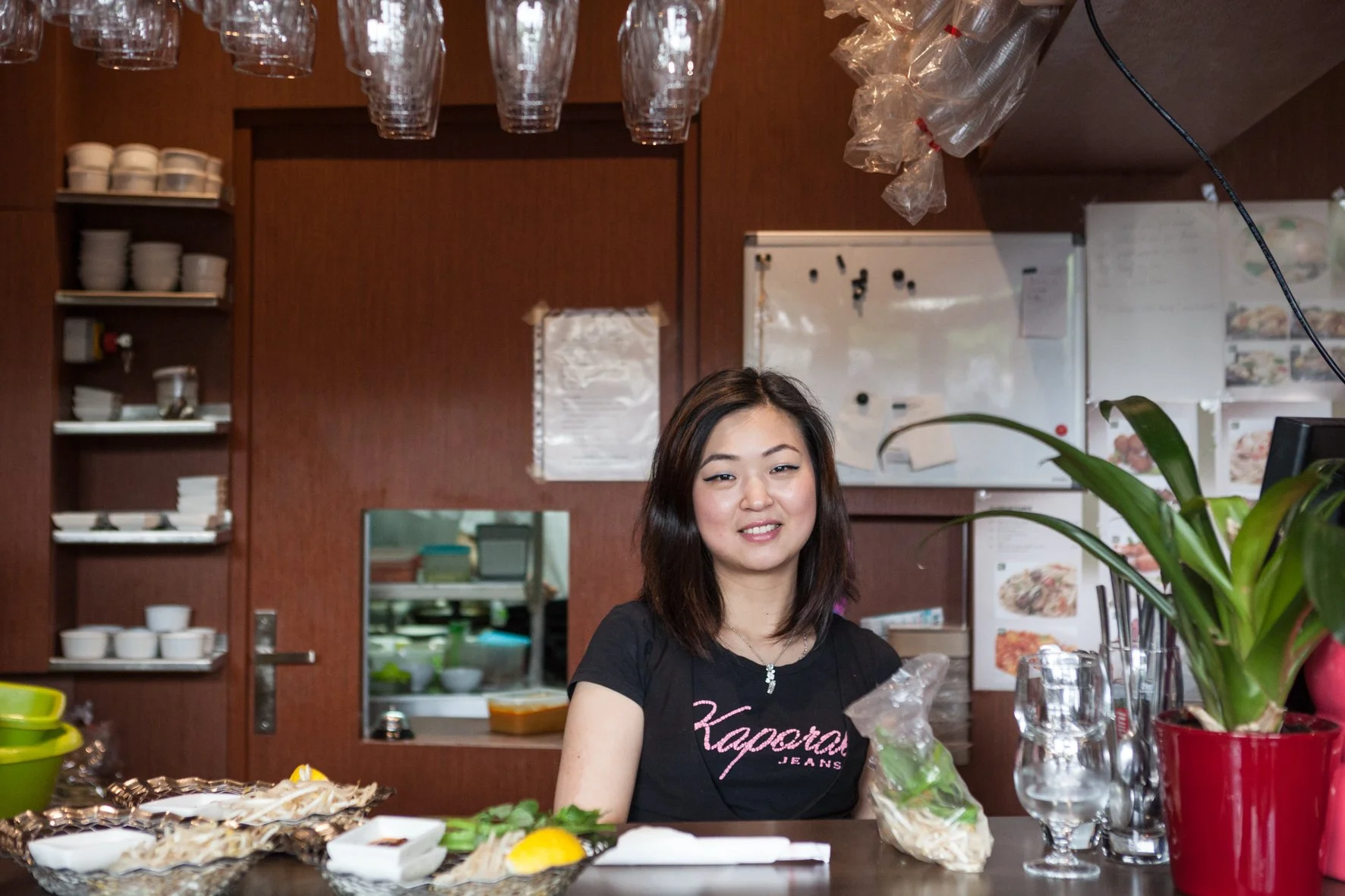 Une femme asiatique souriante derrière un comptoir de restaurant de sushi, entourée de plats, de verres, et de plantes décoratives, avec une kitchenette en arrière-plan.