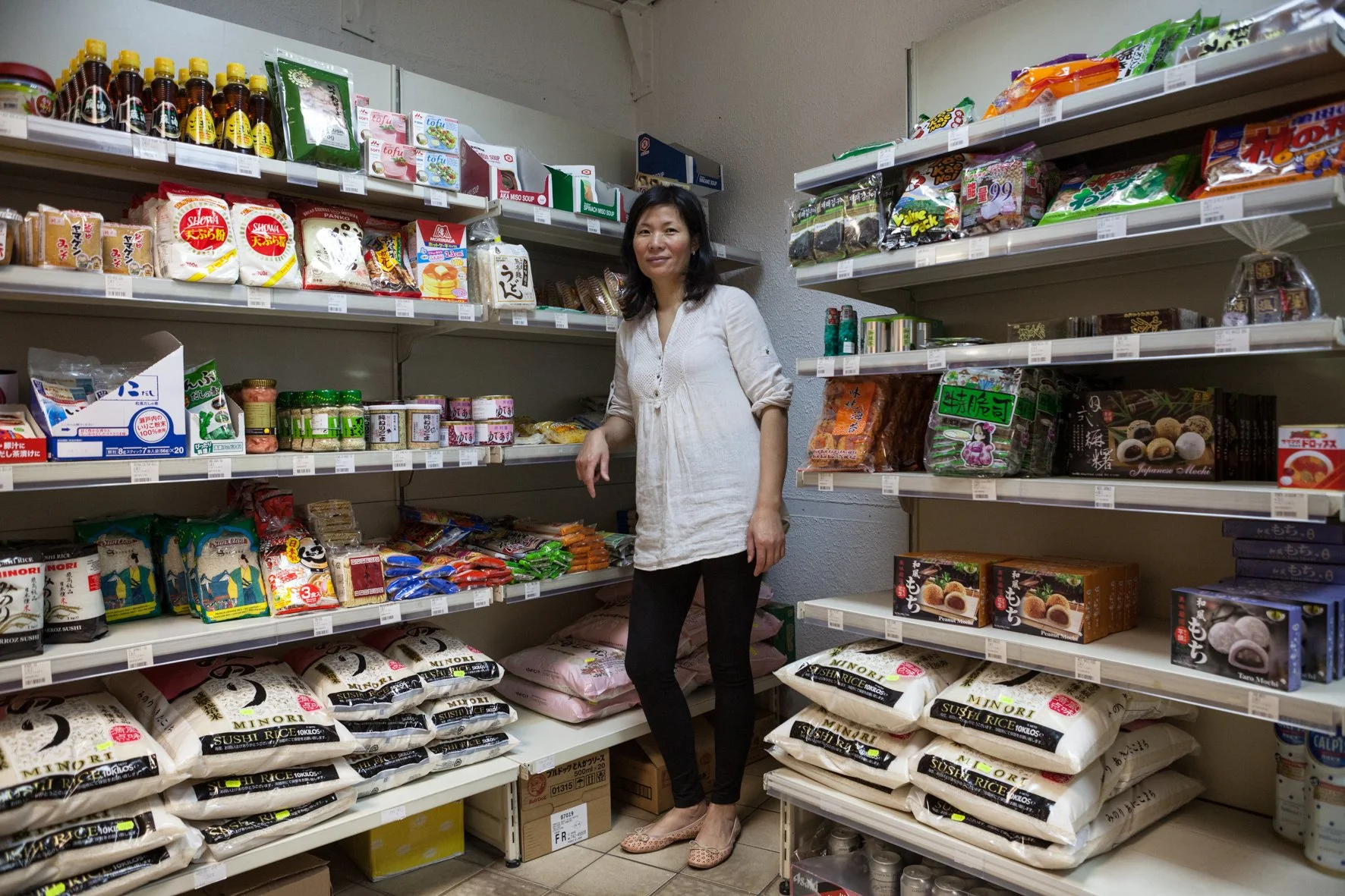 Une femme dans une épicerie ou un rayon de magasin asiatique, entourée de paquets de riz, nouilles, conserves, sauces, snacks et autres produits alimentaires asiatiques.
