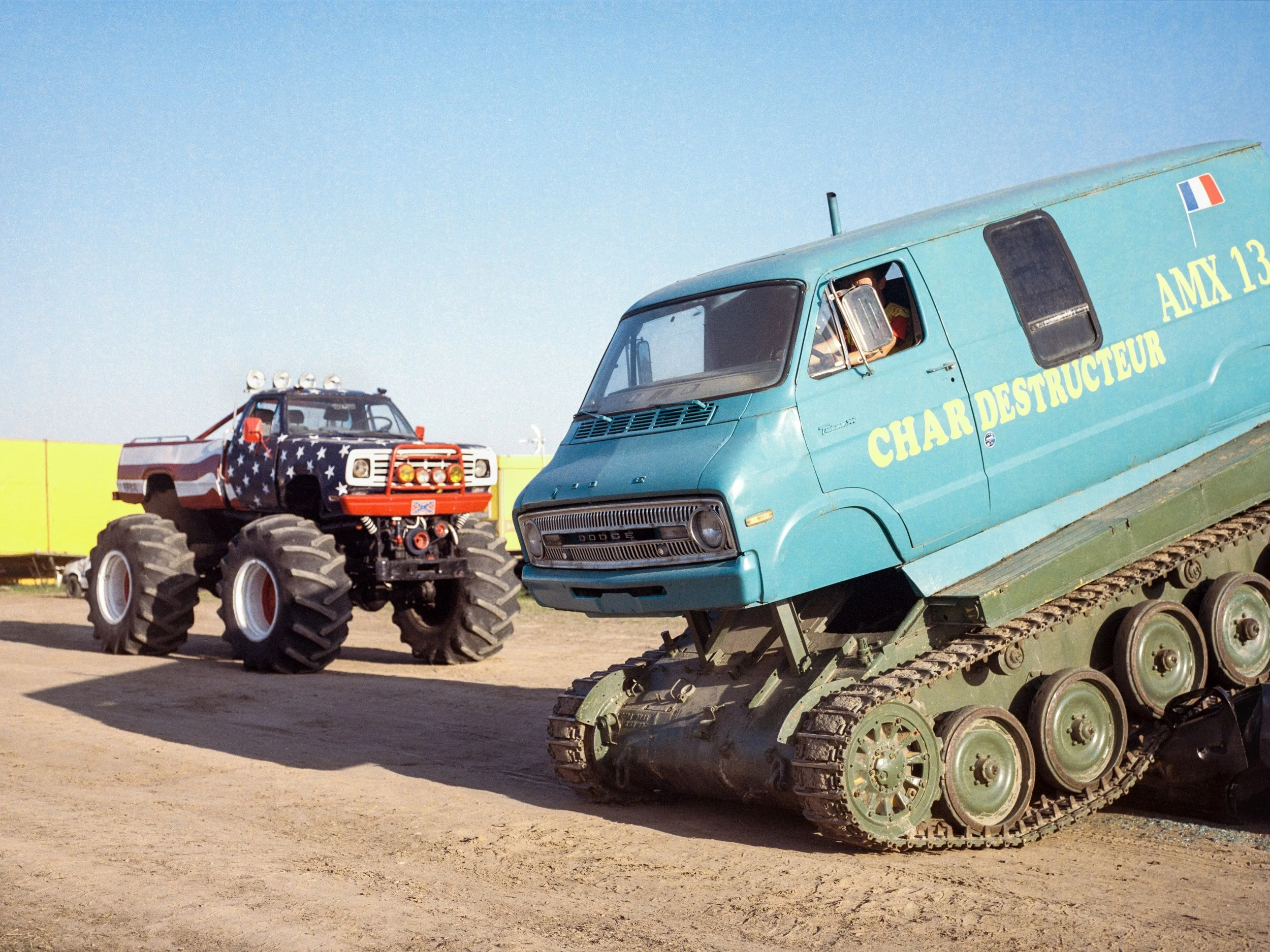 Un véhicule nommé 'Char Destructeur' sur des chenilles, peint en bleu avec l'inscription 'AMX 13' et un drapeau français, stationné à côté d'un autre véhicule de type Monster Truck à grandes roues conçu pour les spectacles.