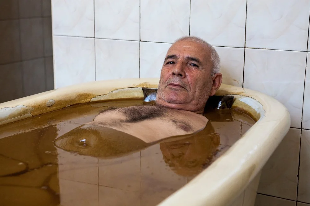 Un homme âgé allongé dans une baignoire remplie d'eau brunâtre, dans une pièce avec des murs en carreaux blancs.