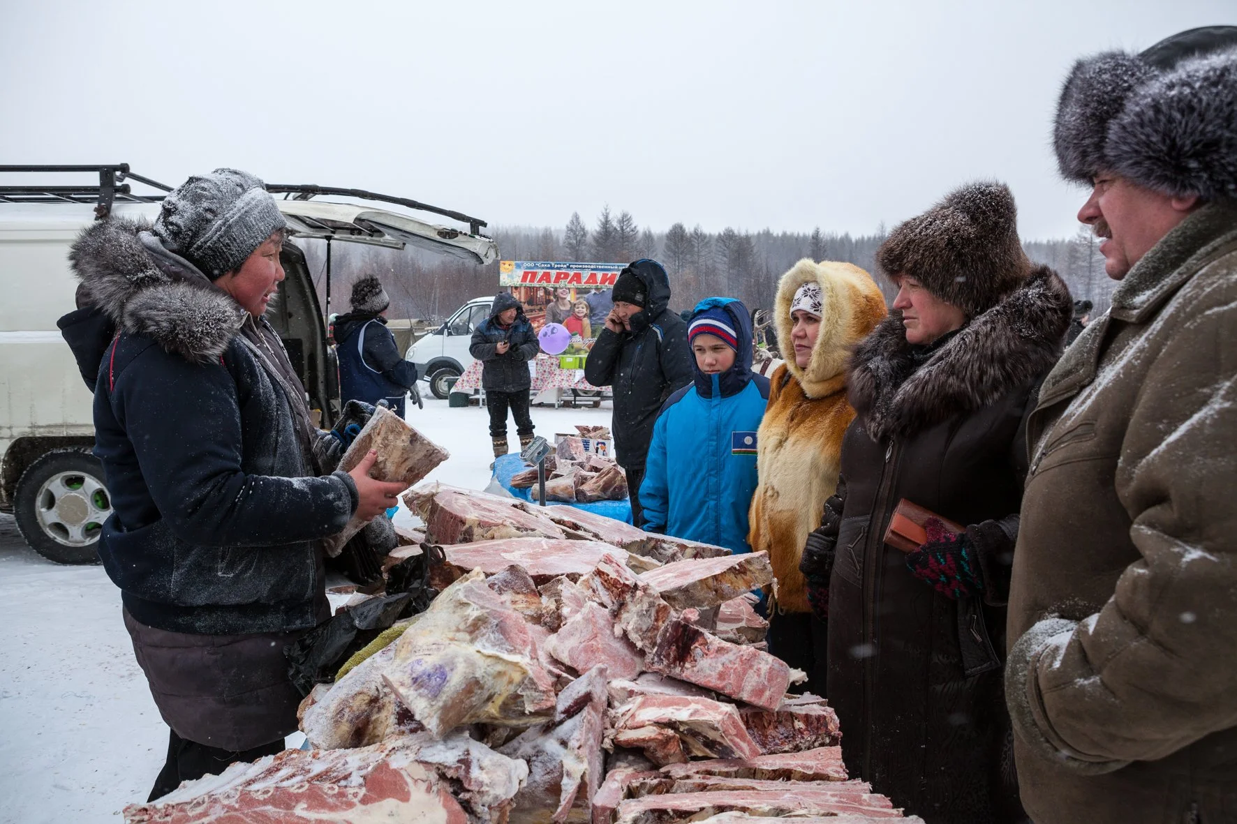 Groupe de personnes achetant de la viande à un stand en plein air en hiver, avec neige et arbres nus en arrière-plan.