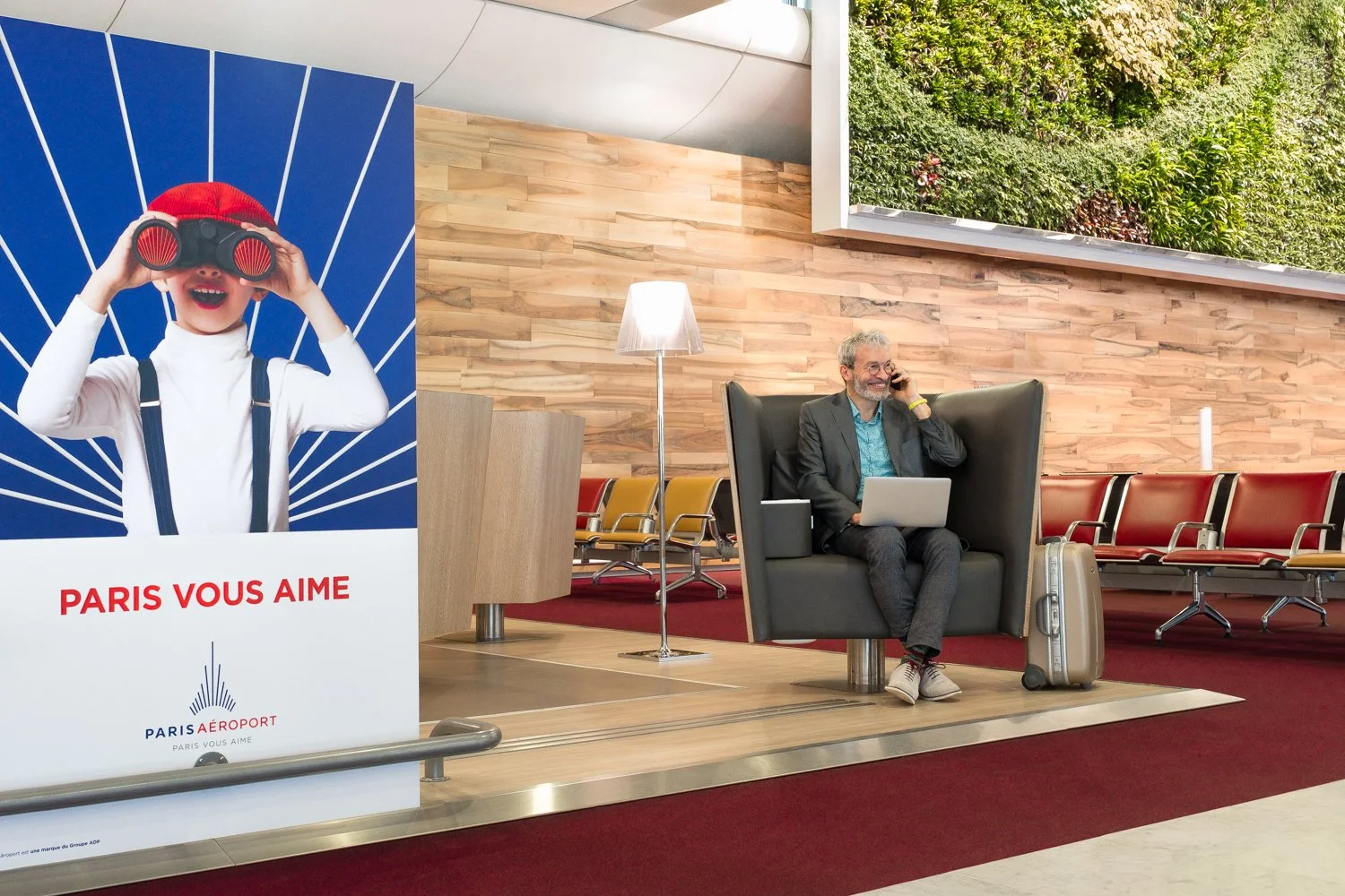 Un homme assis dans une salle d'attente à l'aéroport, parlant au téléphone portable, avec un ordinateur portable sur ses genoux et une valise à ses côtés, derrière une affiche publicitaire de Paris Aeroport représentant un enfant regardant à travers 