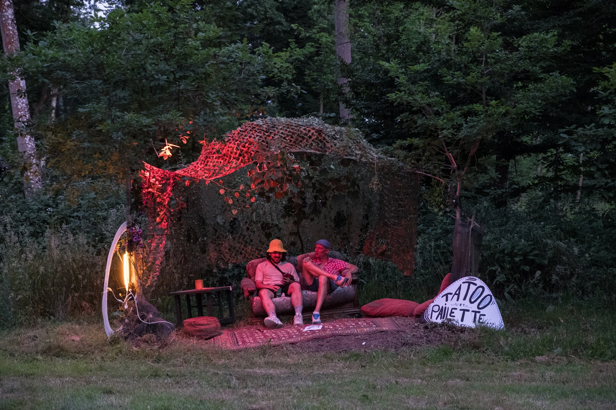 Deux personnes assises sur un canapé en plein air, sous un camouflage fait de branches et de feuilles, entourées de coussins, avec une petite table à côté.