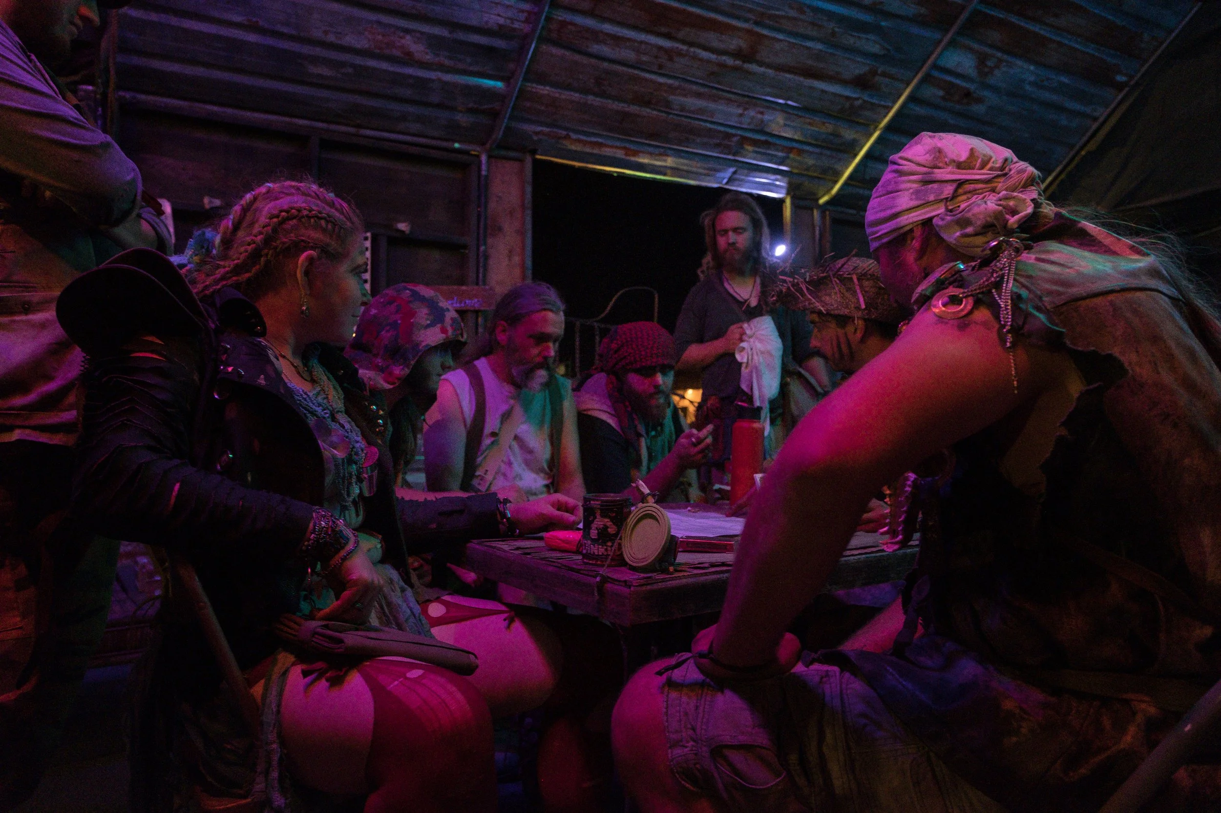Groupe de personnes assises autour d'une table dans une ambiance sombre et colorée, discutant ou participant à une activité à Wasteland.