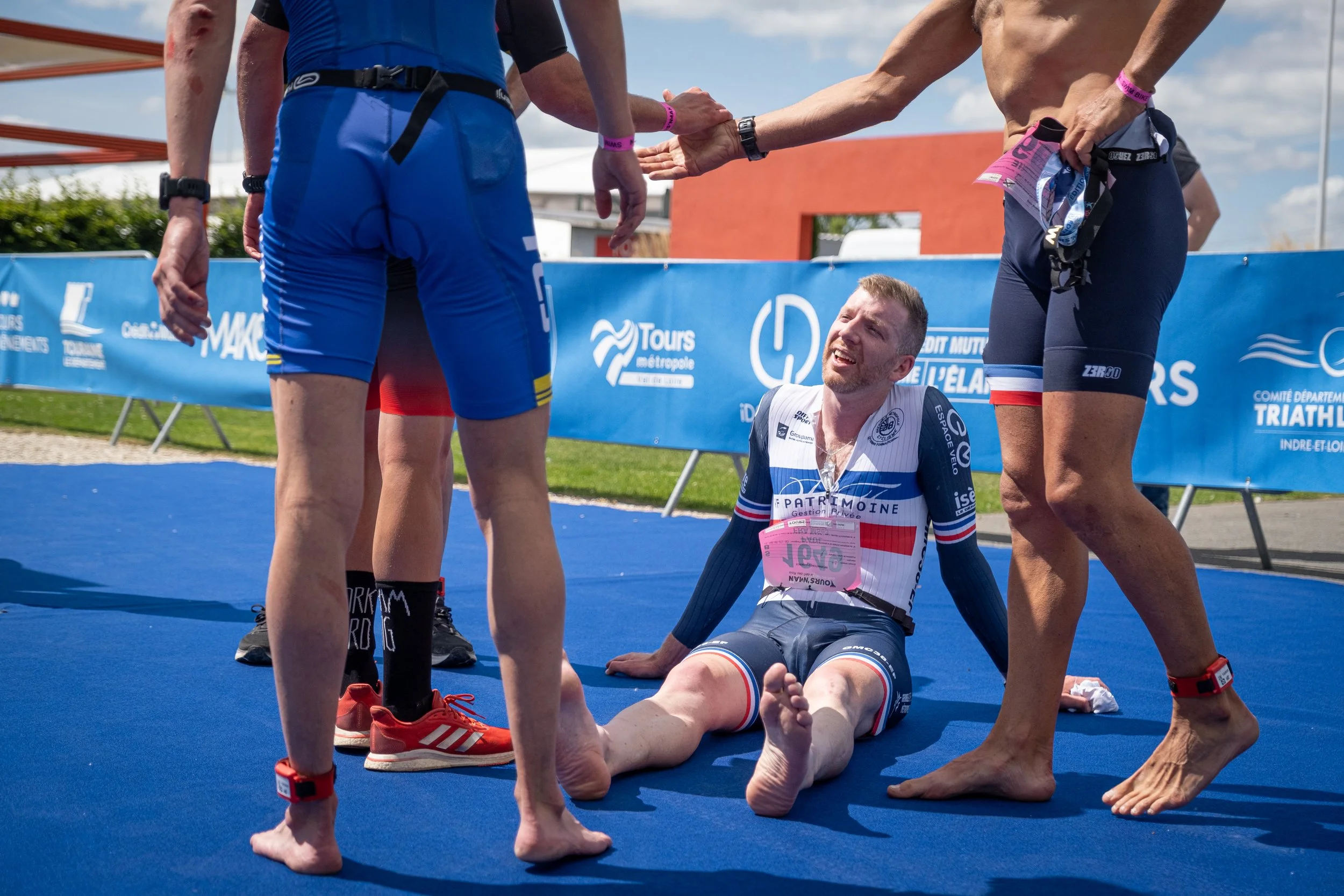 Un athlète en tenue de triathlon assis sur le sol, visiblement épuisé, entouré de plusieurs autres athlètes qui lui tendent la main pour l'aider à se relever, en plein événement sportif en plein air.