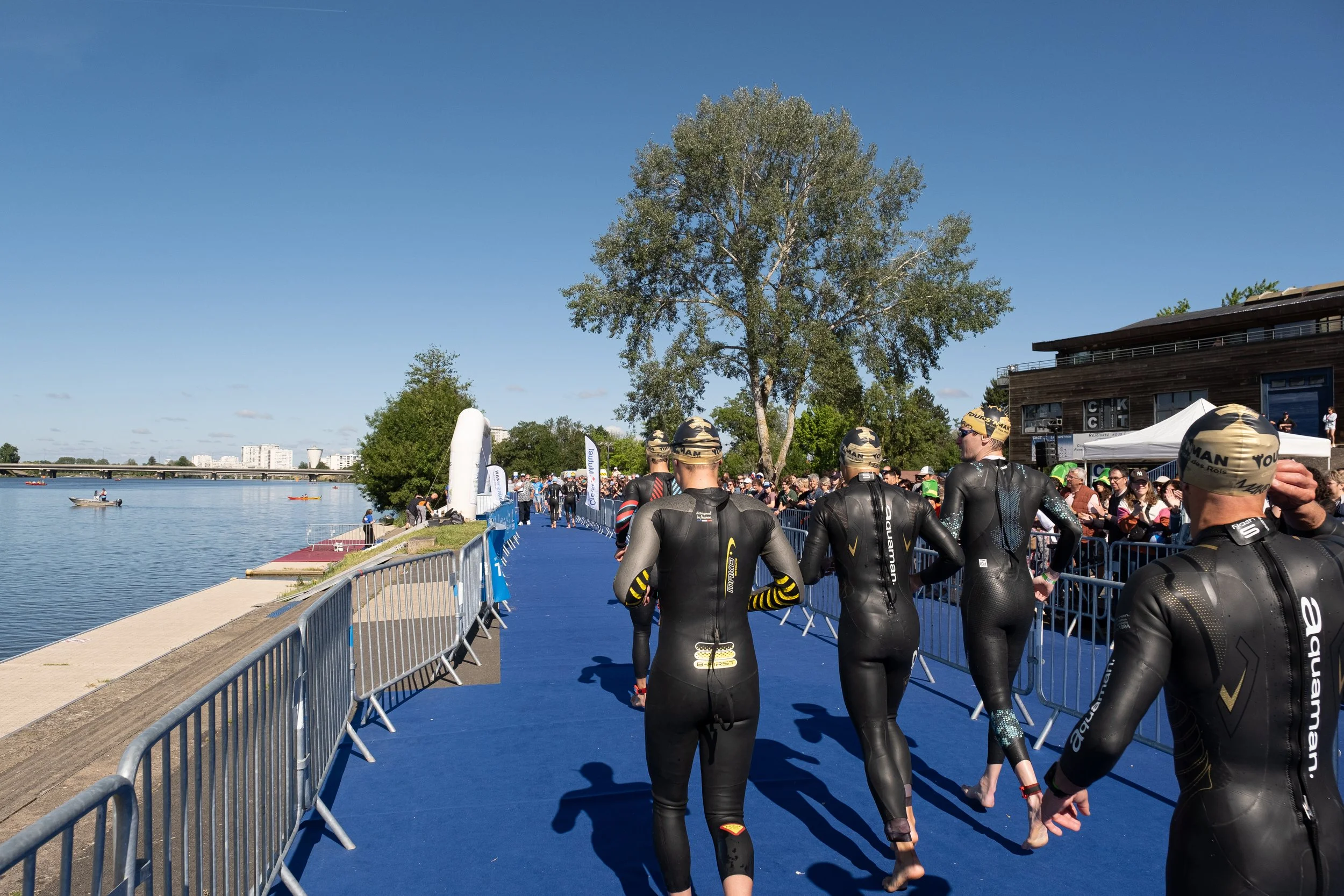 Des nageurs en combinaison de triathlon marchent sur une passerelle bleue près d'une rivière lors d'une compétition, avec un public et des bénévoles en arrière-plan.